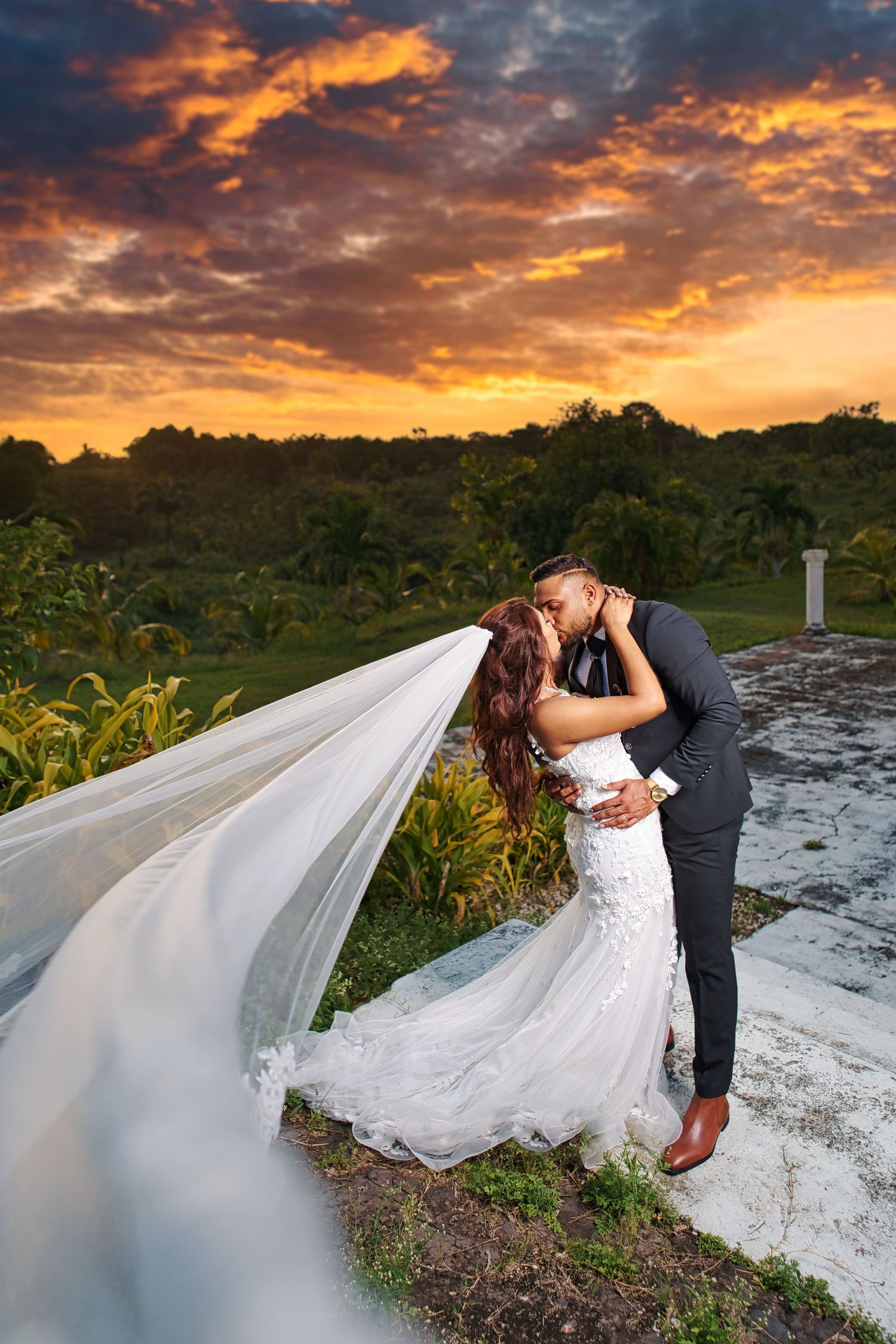 A couple in traditional South Asian wedding attire embraces under palm trees in a tropical garden setting.
