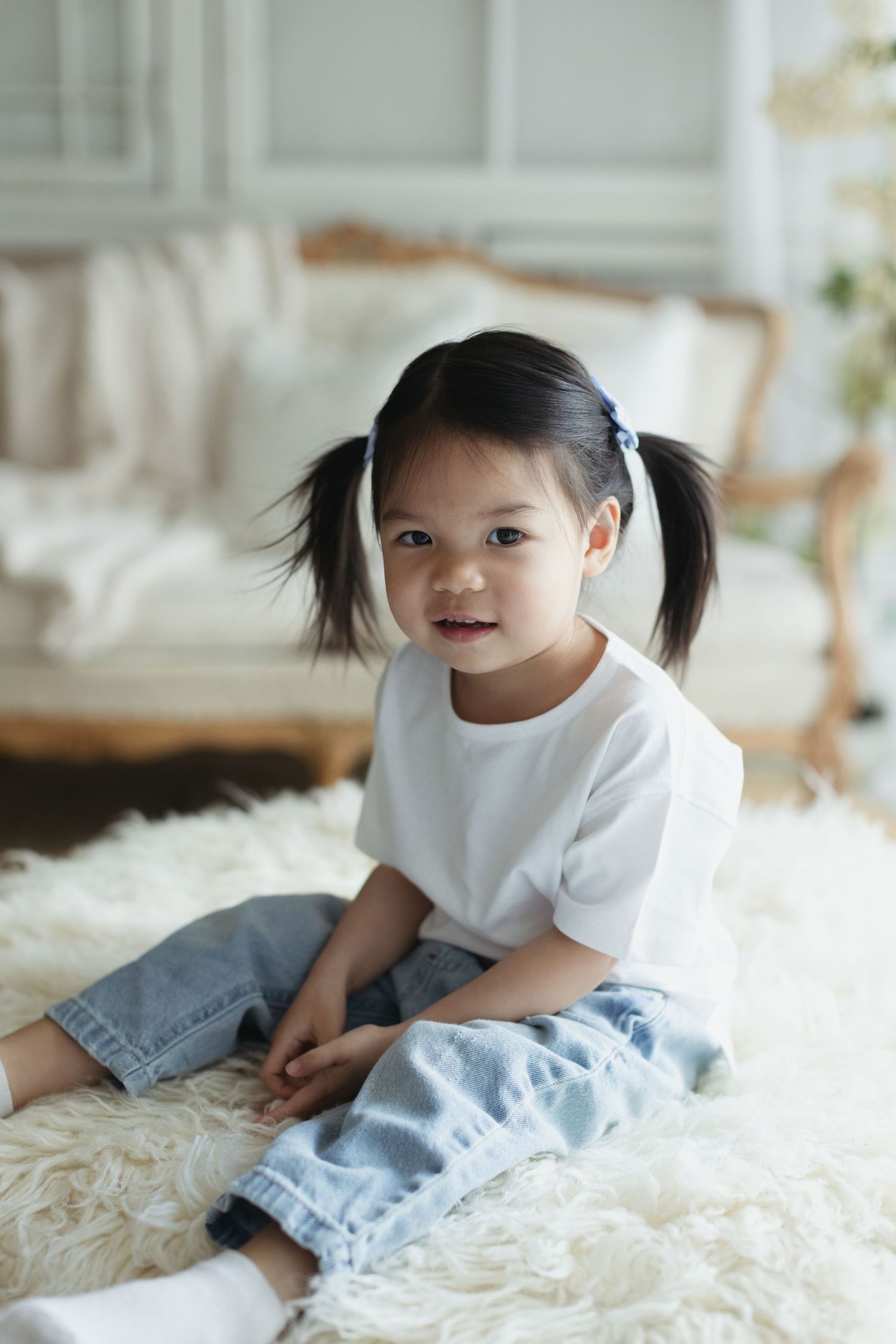 Cute Asian toddler girl with pigtails sitting on fluffy white rug in white t-shirt and jeans.