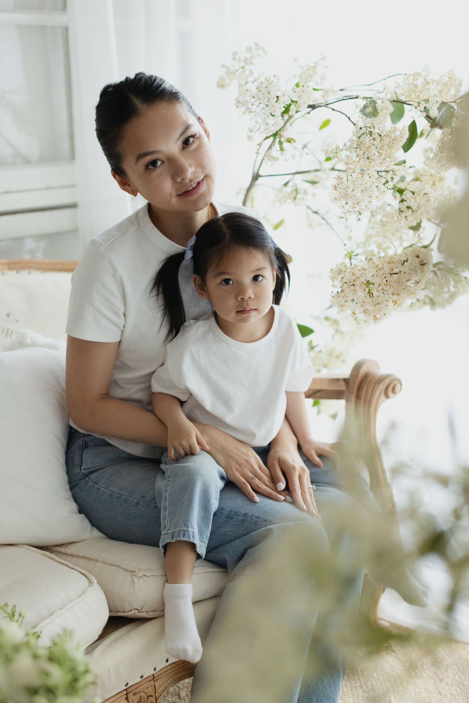 Asian mother and daughter in matching white tees and jeans sitting together on a couch surrounded by white flowers.