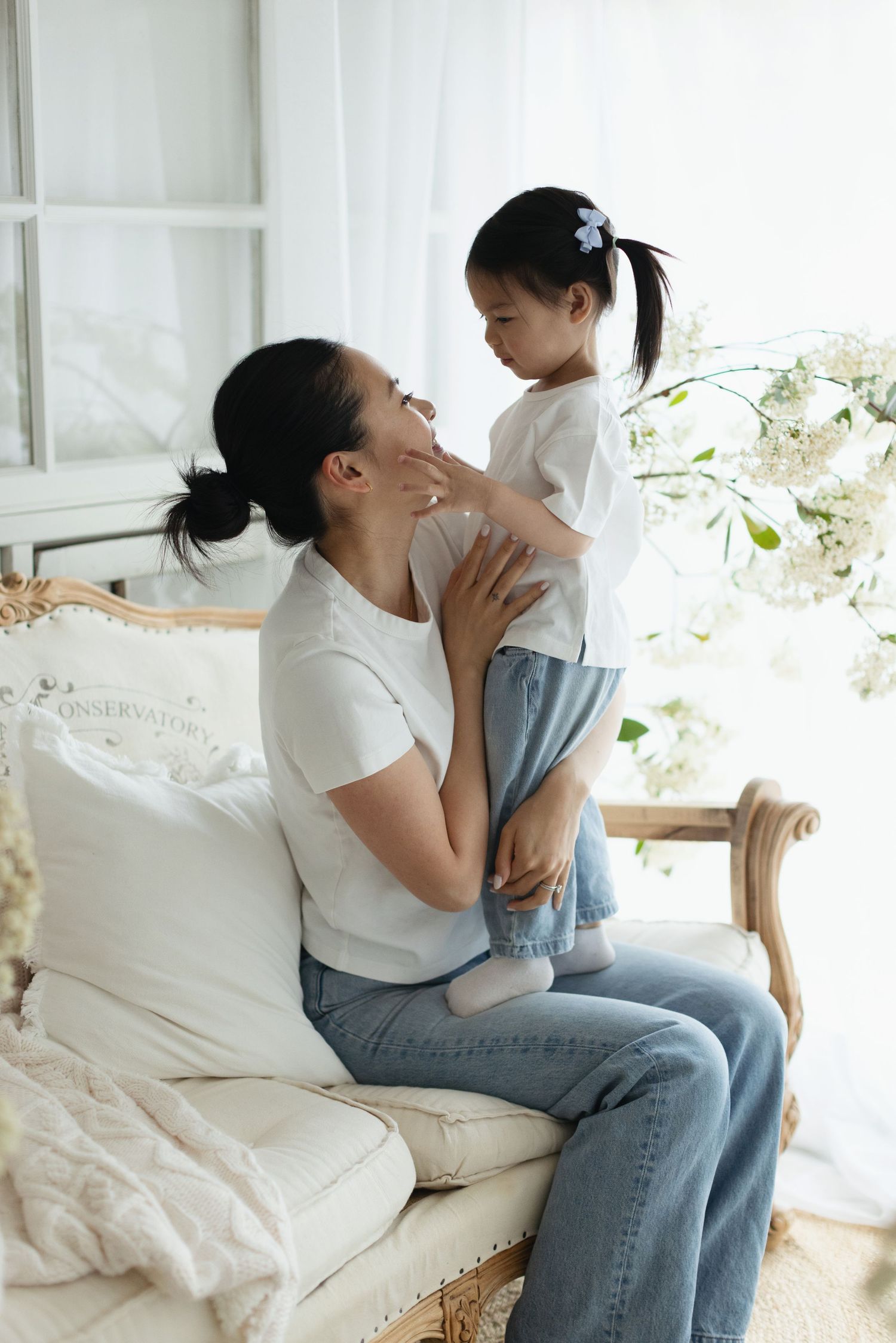 A mother sits on an elegant chair holding her young daughter, sharing a tender moment in a bright, white-decorated room.
