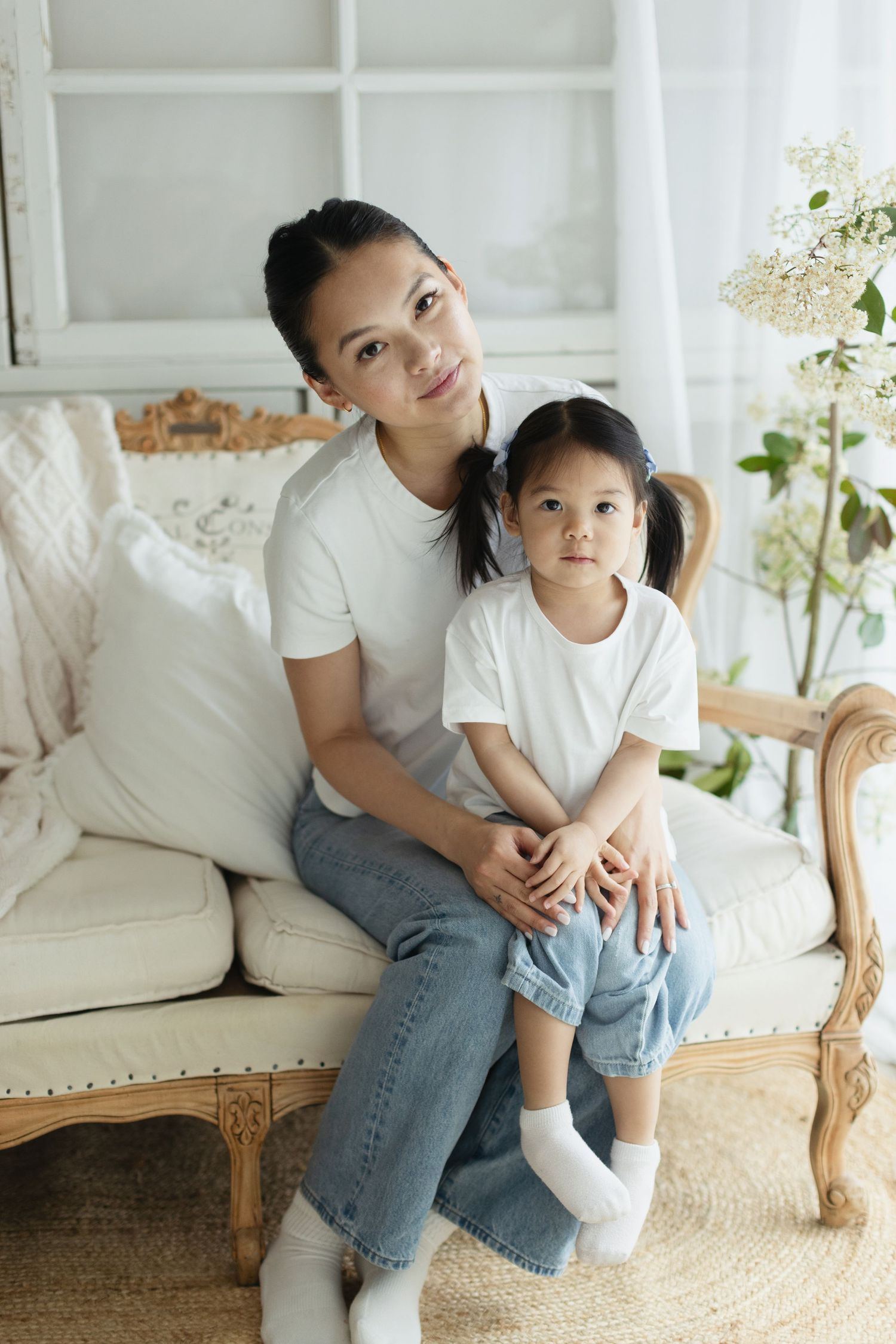 Asian mother and daughter in matching white tees and jeans sitting together on a vintage sofa at home.