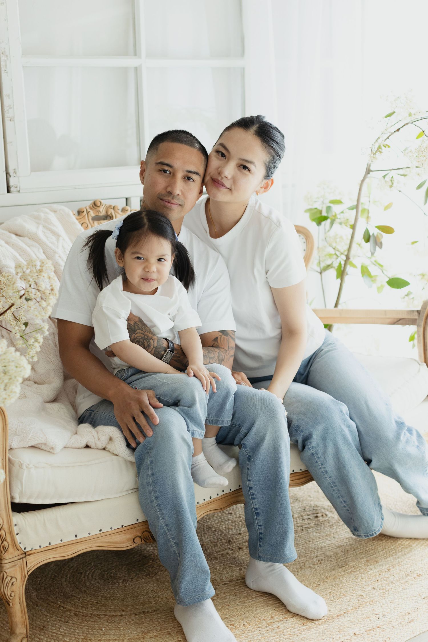 Family of three in matching white tees and jeans posing on a vintage sofa in a bright, airy room.