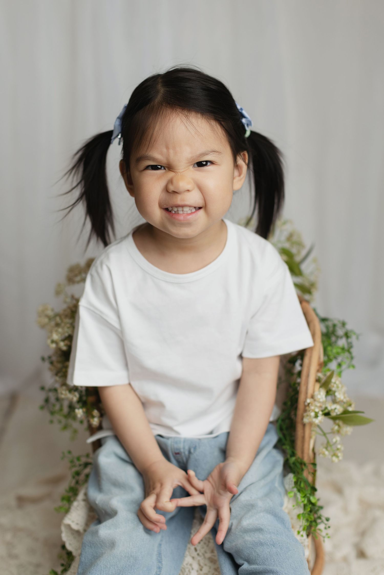 Adorable Asian little girl with pigtails making funny face sitting on chair with floral wreath.