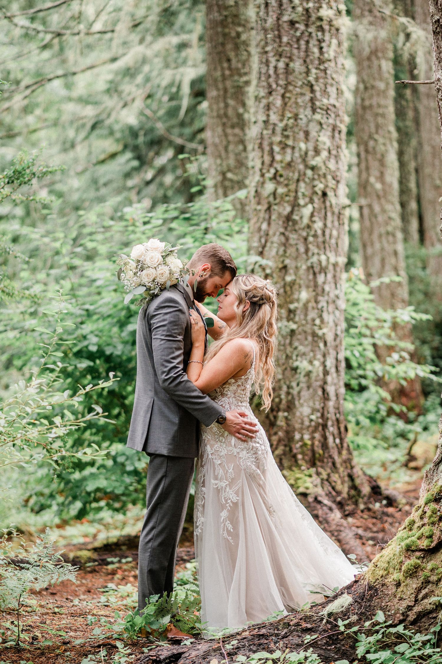 Bride and groom share a romantic kiss in a lush forest setting, with the bride holding a white bouquet.