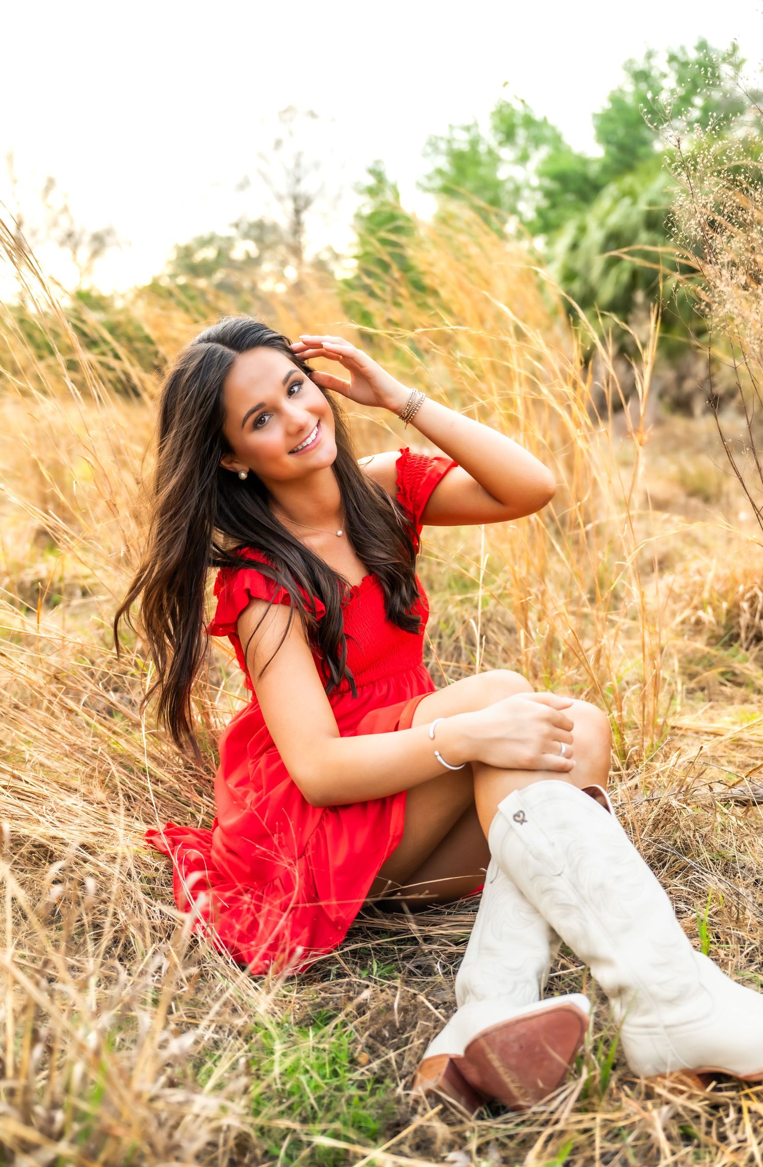 Young woman in red dress and white cowboy boots sitting in golden field grass, smiling outdoors.