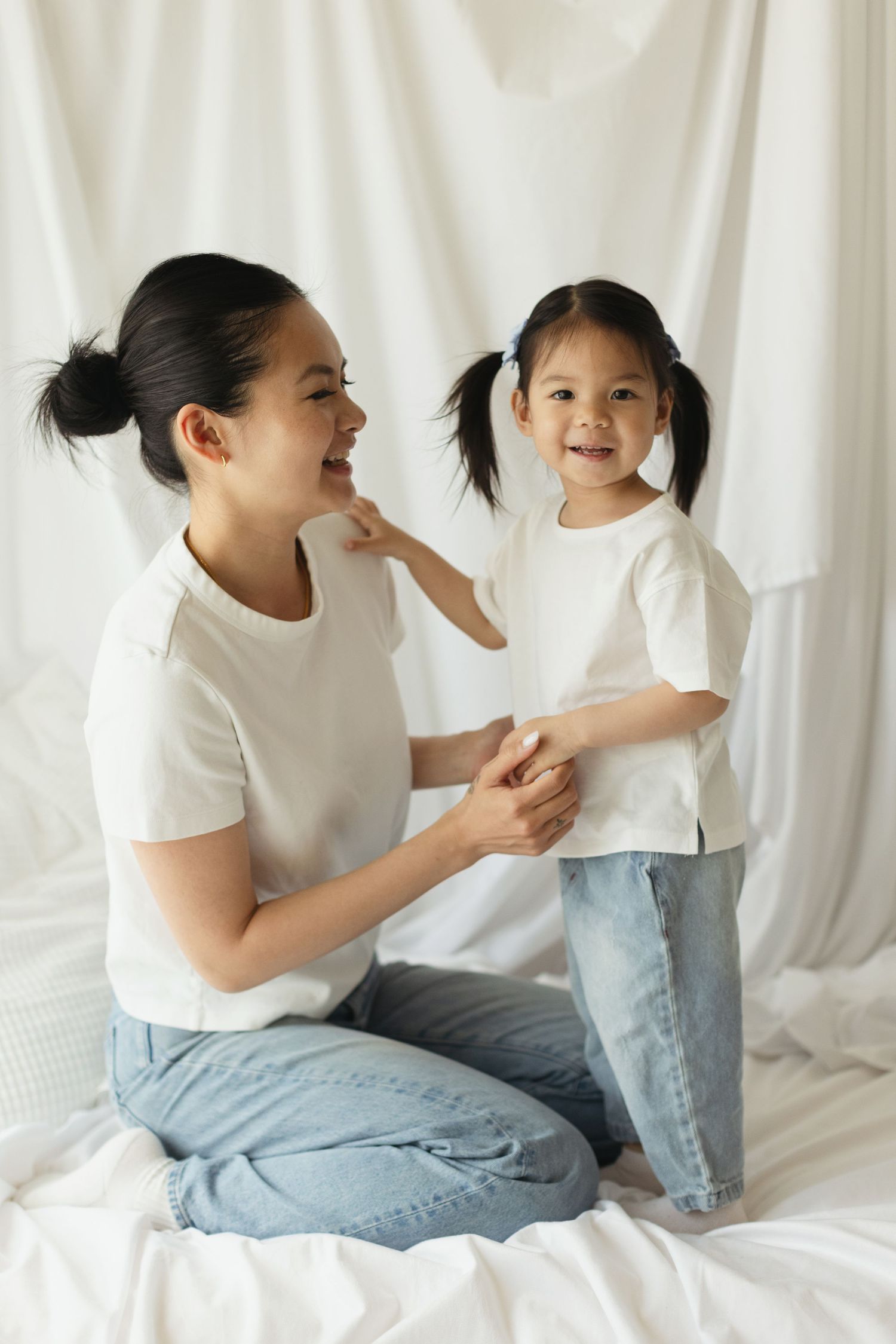 Asian mother and young daughter in matching white tees and jeans sharing a joyful moment on a white bed.