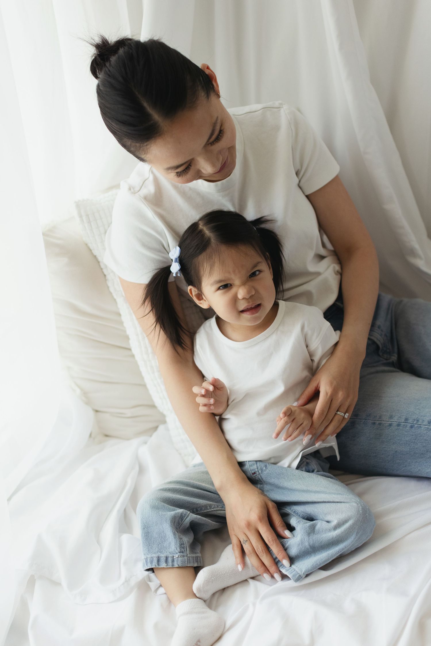 Asian mother and young daughter in matching white tees and jeans sitting together on a white bed.