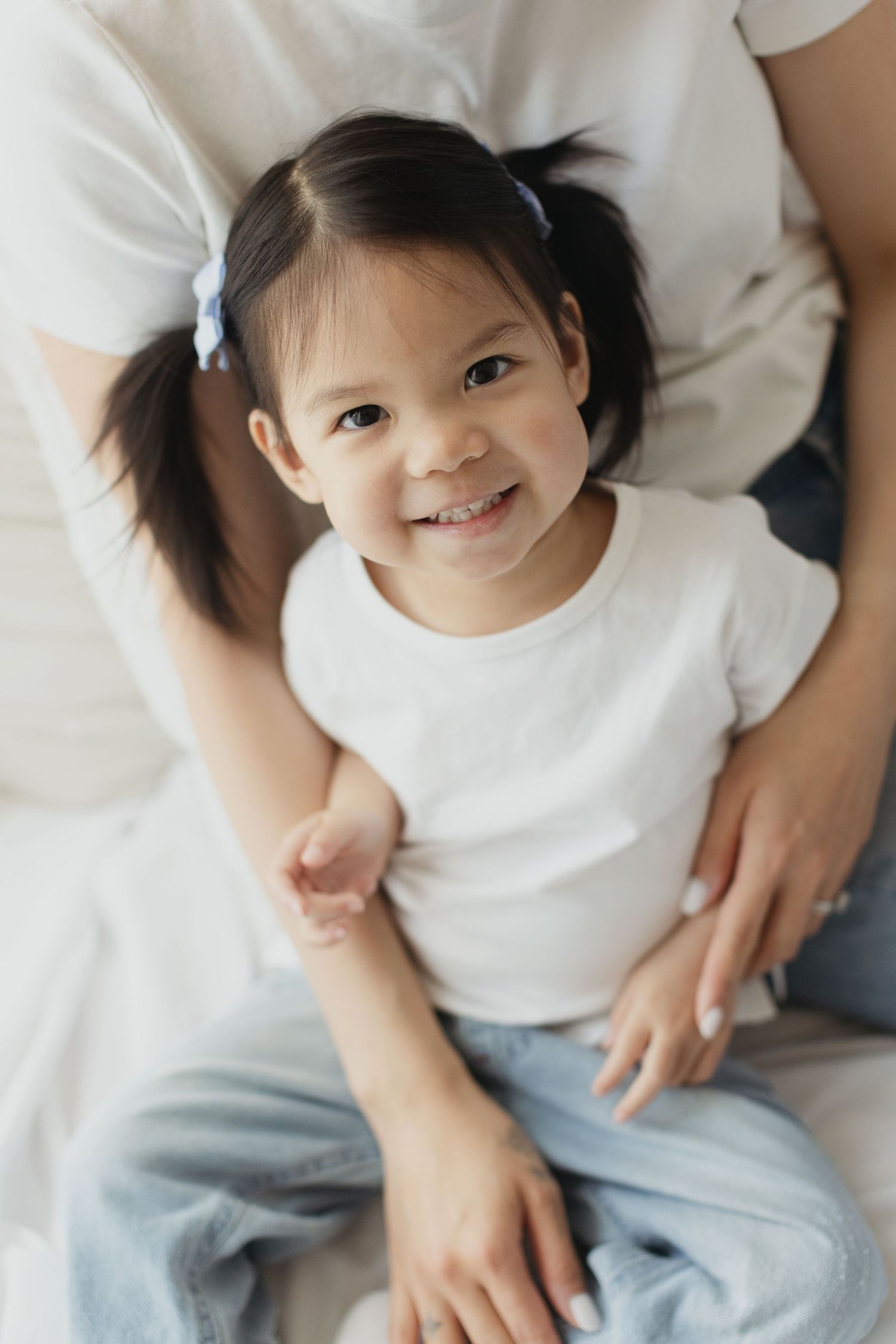 A smiling young Asian girl with pigtails in a white t-shirt and jeans sits in front of an adult on a bed.