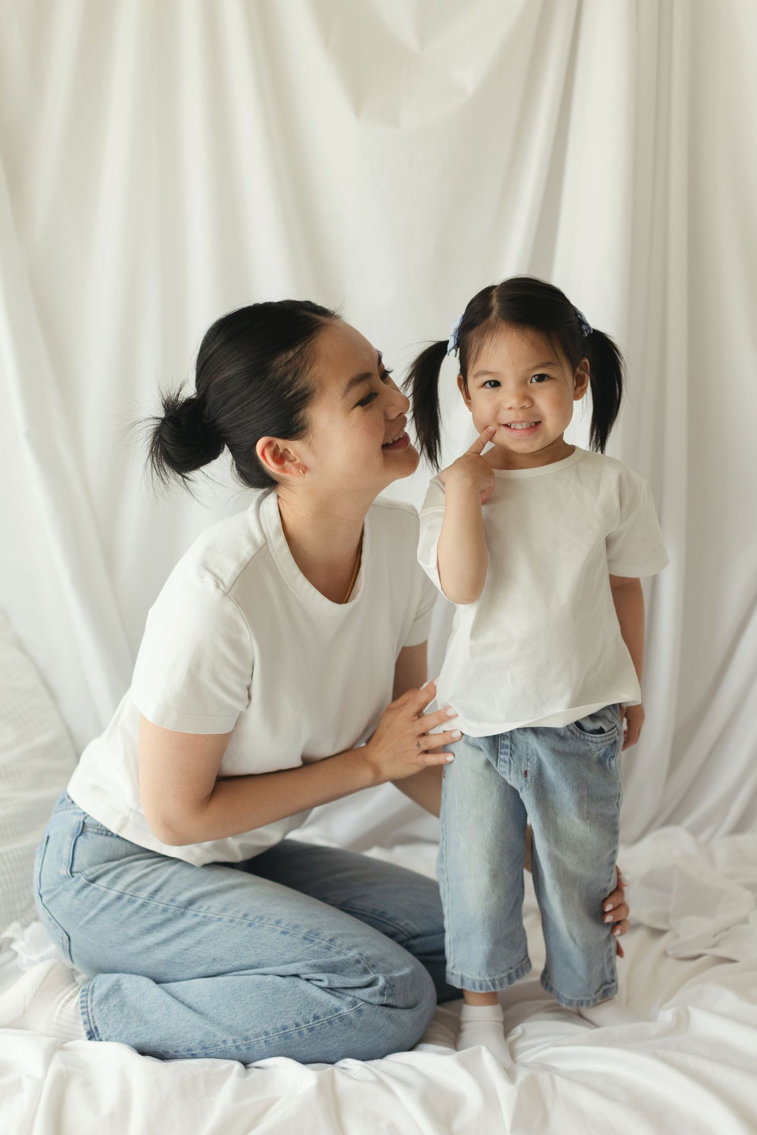 Asian mother in white tee and jeans kneeling beside smiling toddler daughter with pigtails against white backdrop.