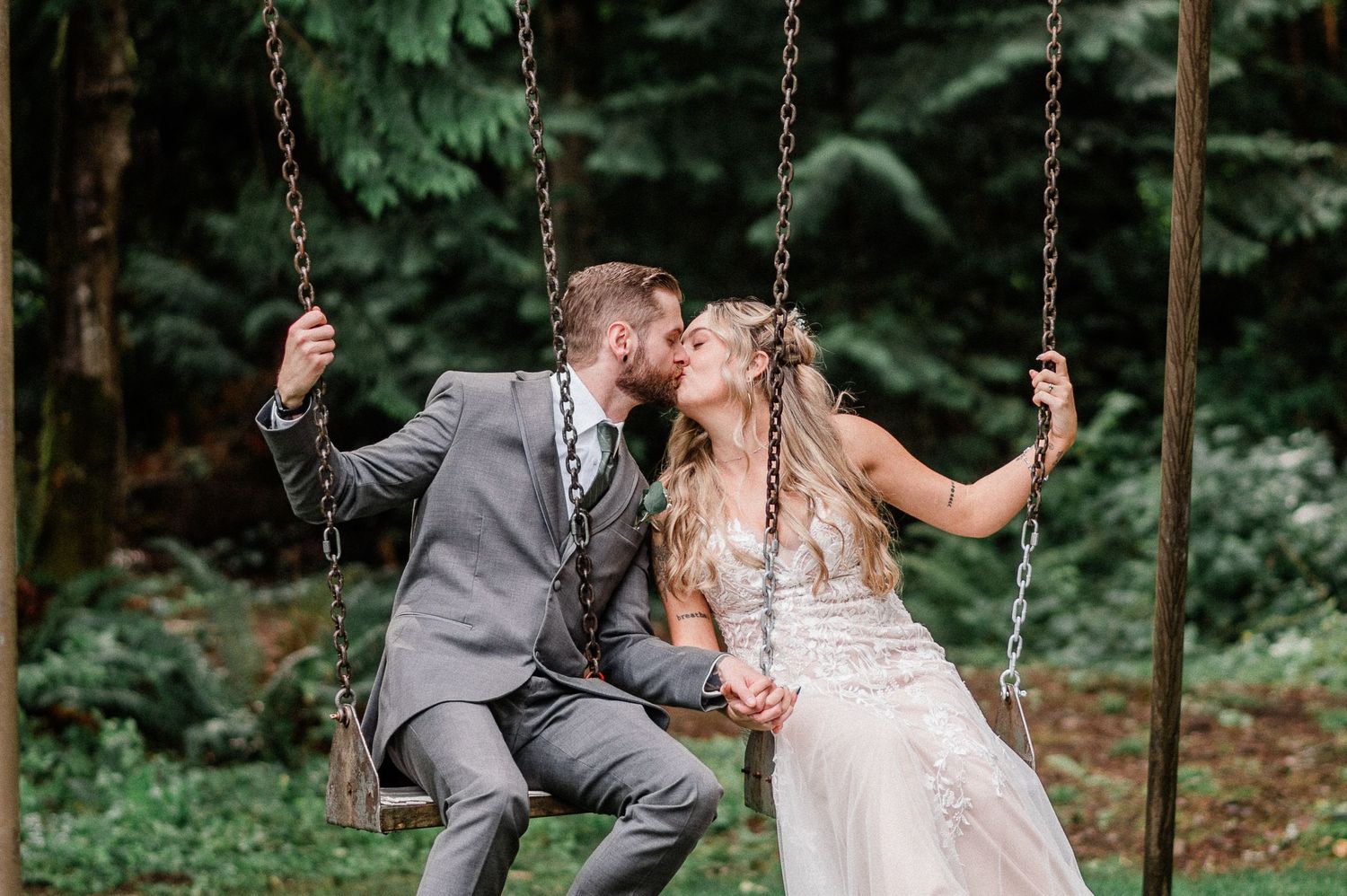 Bride and groom kissing on a wooden swing surrounded by lush green forest on their wedding day.
