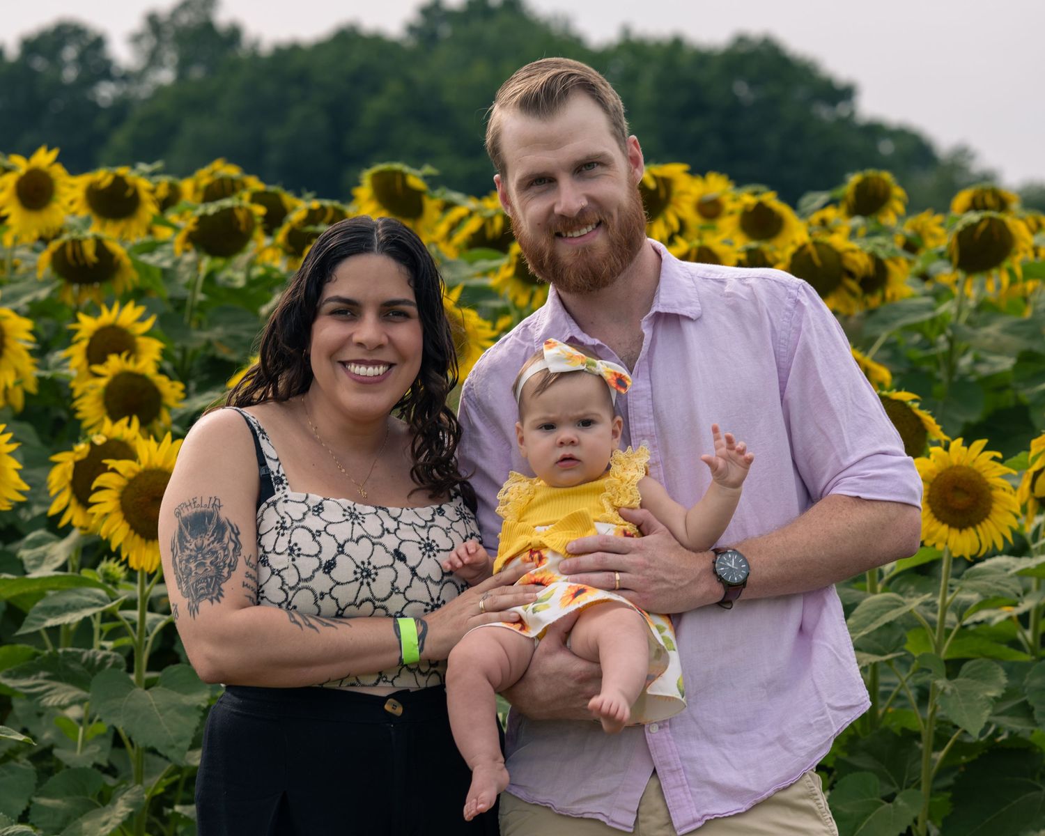 Young couple holding their baby girl in a yellow bow headband in a beautiful sunflower field.