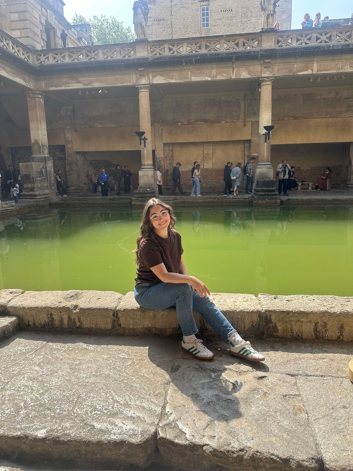 Young woman sitting on stone edge of Roman Baths with vivid green thermal pool and ancient architecture in Bath, England.