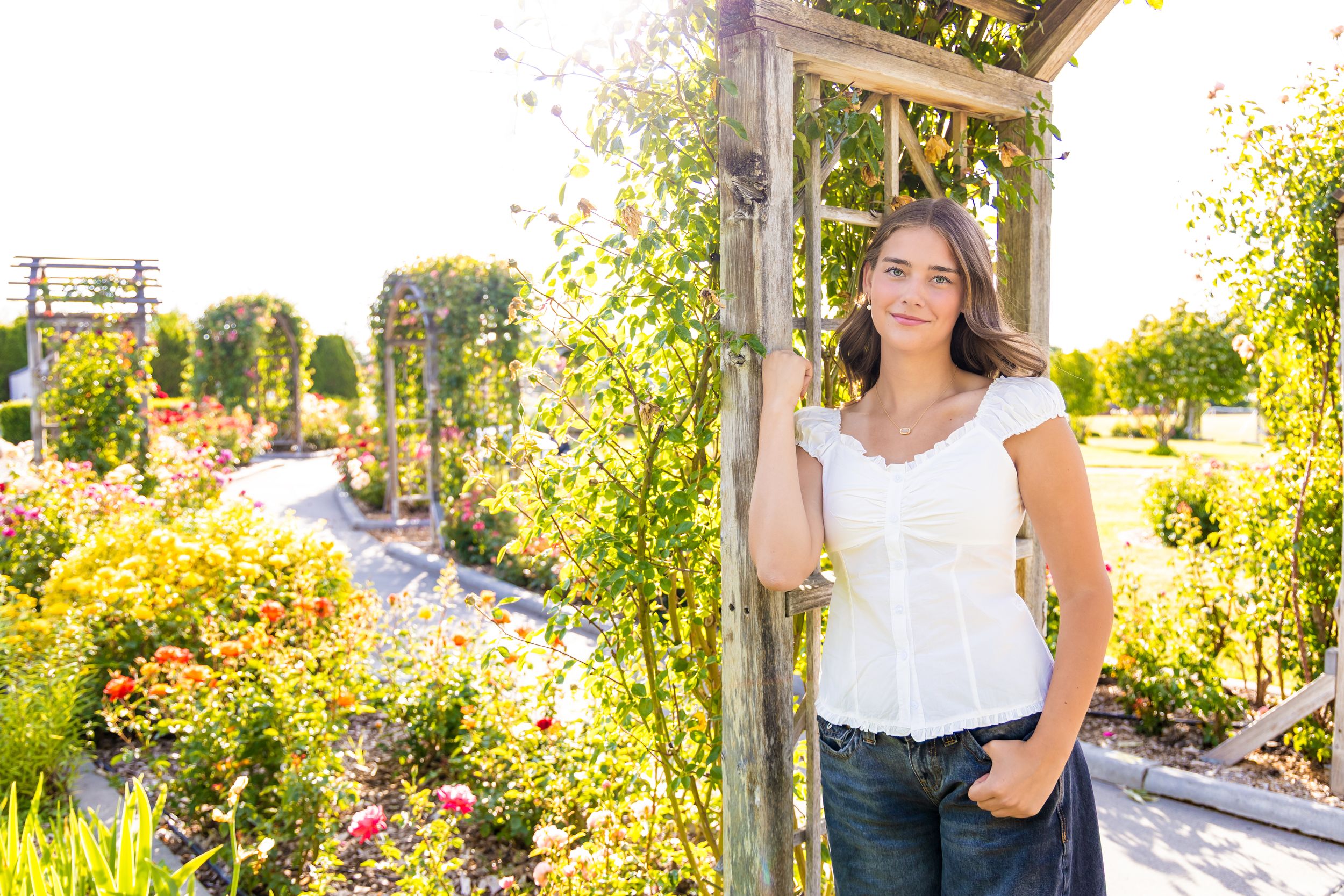 Young woman in white top and jeans posing by a wooden garden arbor surrounded by colorful flowers and lush greenery.
