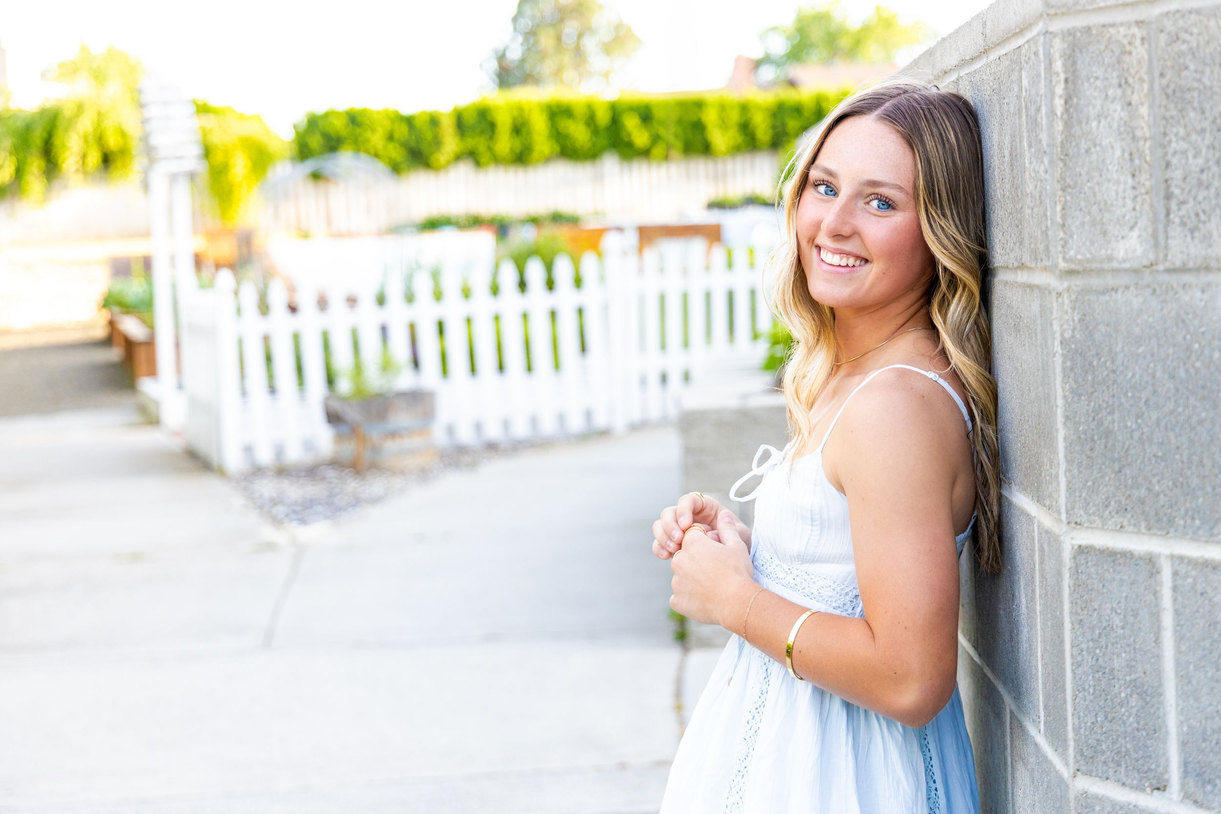 Smiling young woman in a white dress leaning against a gray brick wall outdoors near a white picket fence.