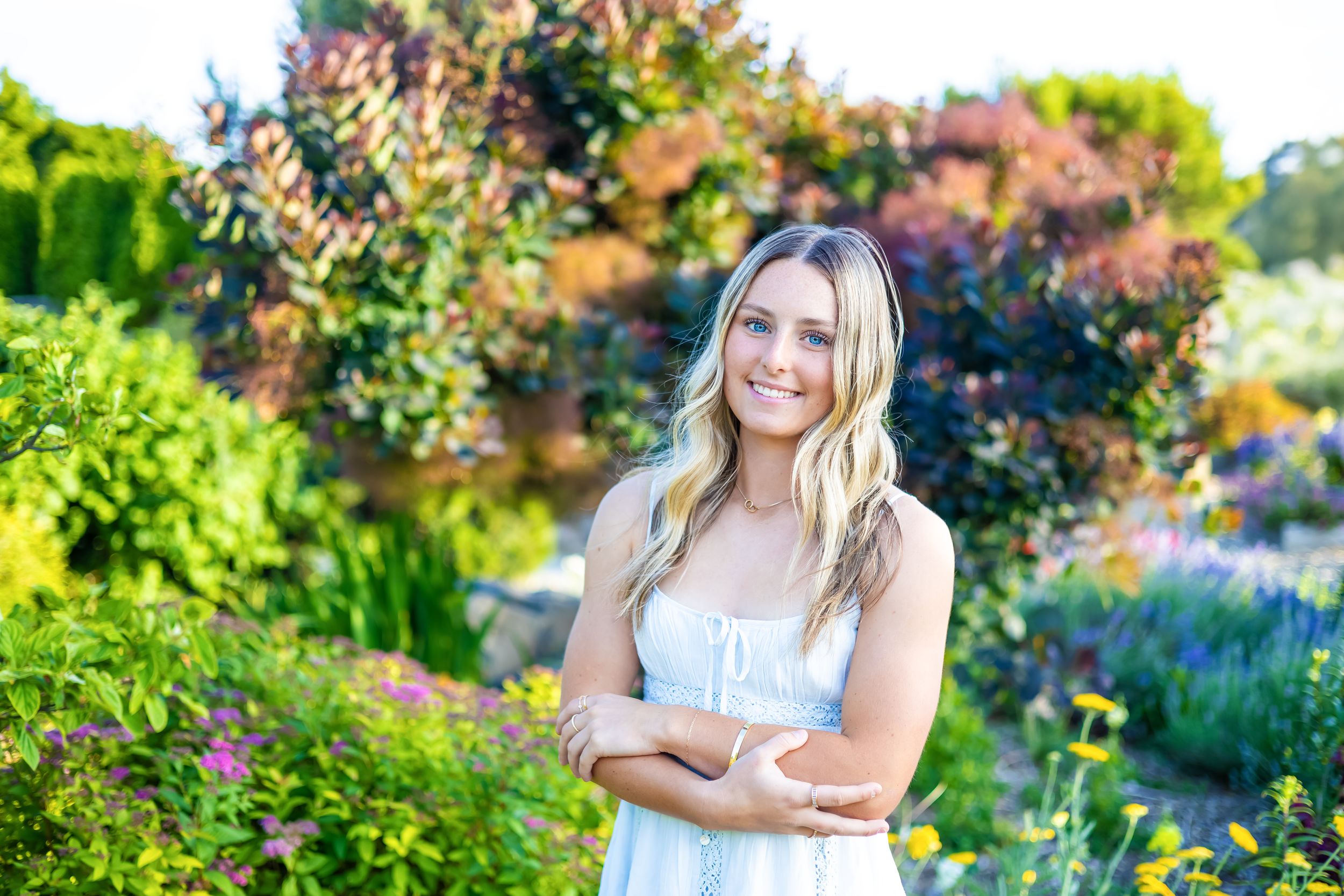 Smiling young woman in white dress with arms crossed standing in a vibrant garden with colorful flowers and greenery.