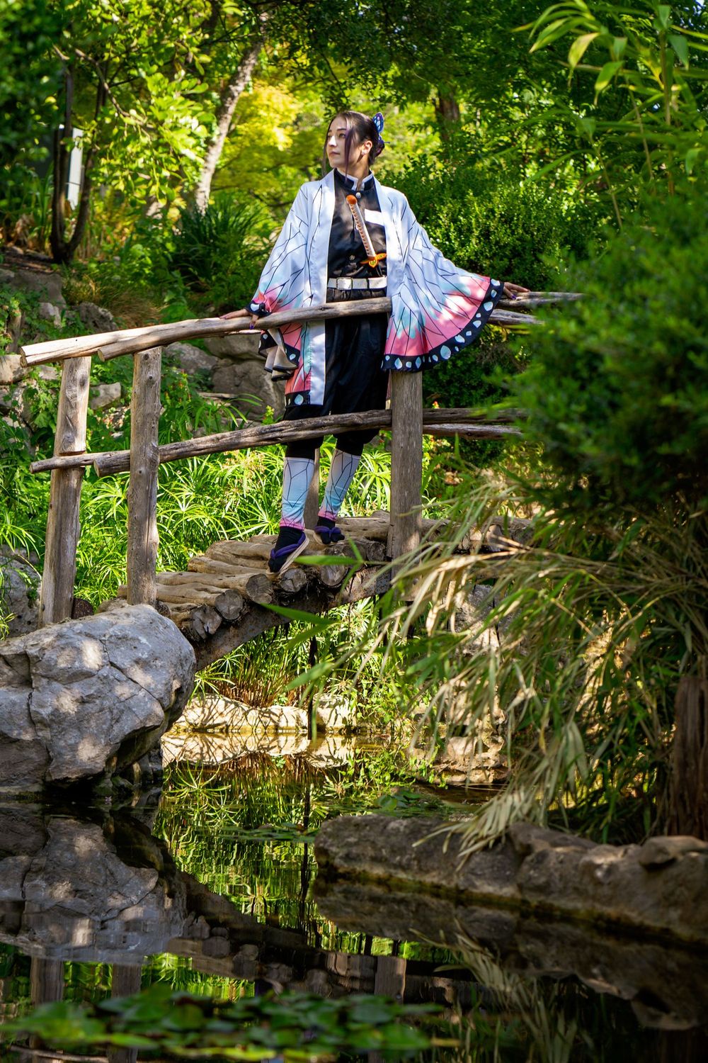 Person in Japanese cosplay costume standing on wooden bridge surrounded by lush green garden with pond reflection.