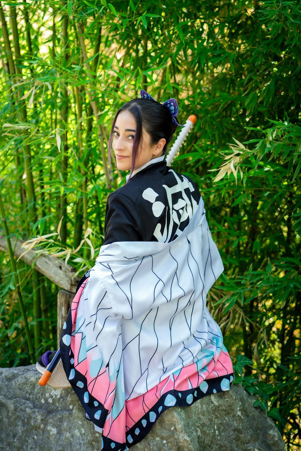 Woman in Nezuko Kamado cosplay costume sitting on rocks in bamboo forest, wearing traditional Japanese kimono.