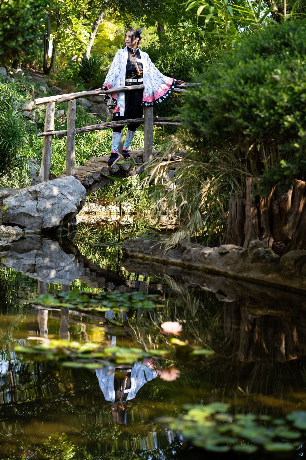 Person in traditional Japanese cosplay costume standing on wooden bridge reflected in lily pad pond.