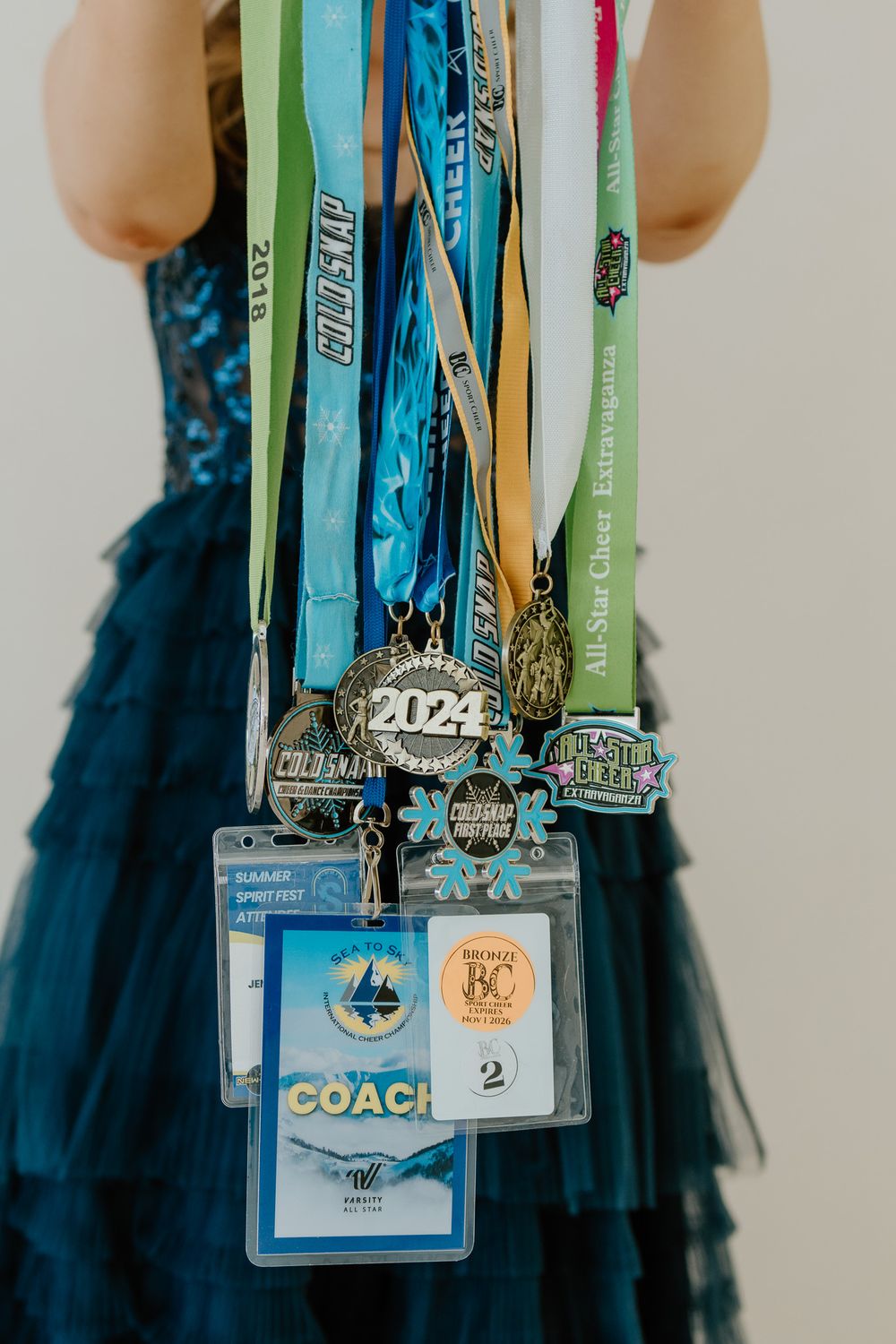 Close-up of cheerleading medals, lanyards, and a coach badge held by a girl in a navy gown.