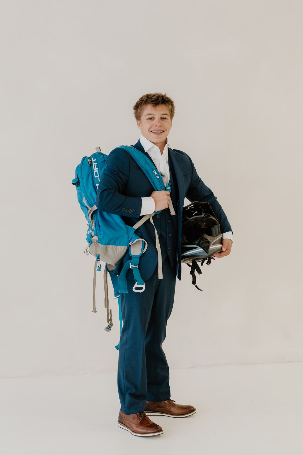 Smiling teen boy in navy three-piece suit holding motocross helmet and teal BCA backpack.