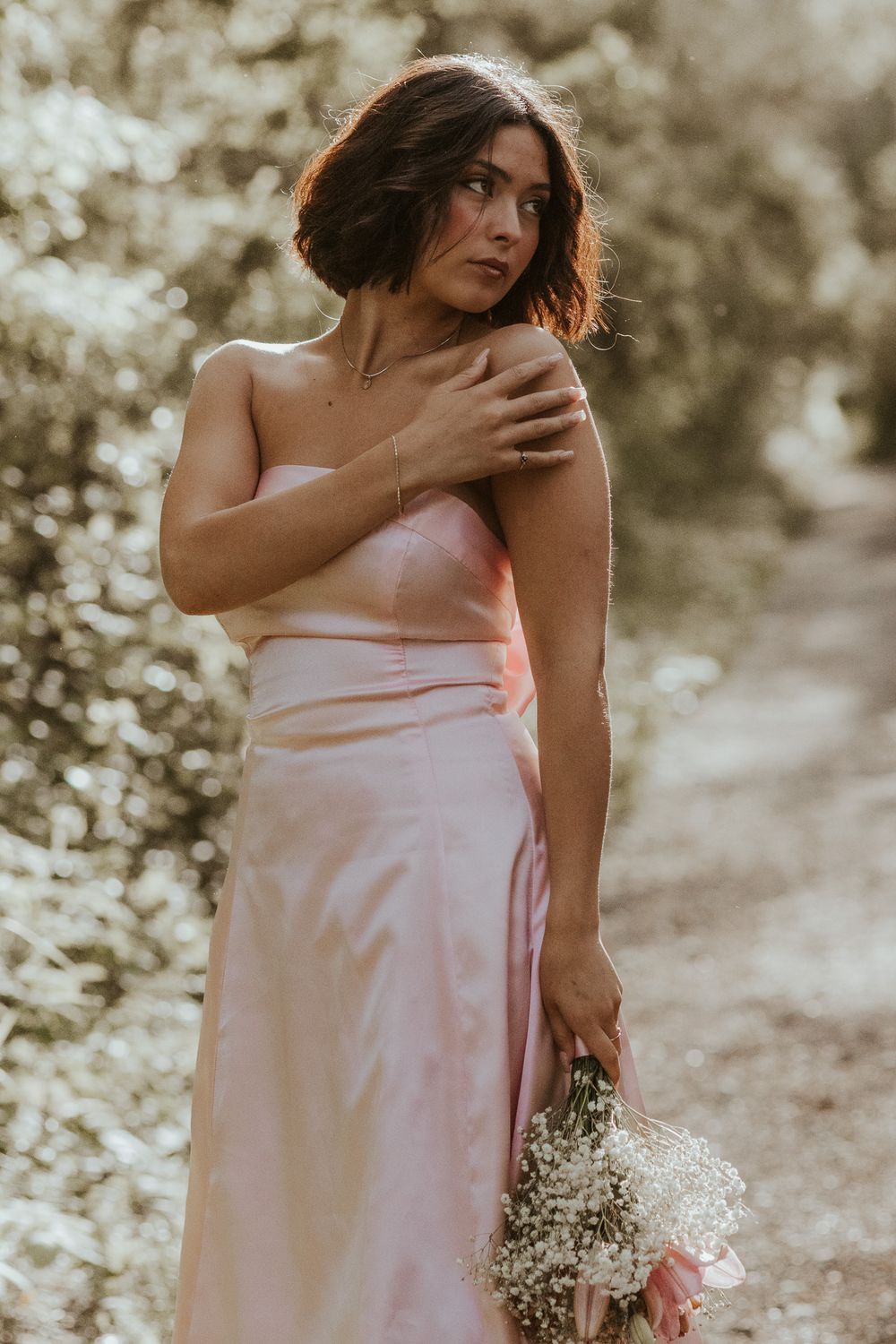 A woman in a pink strapless dress holding flowers, posing gracefully in a sunlit garden.