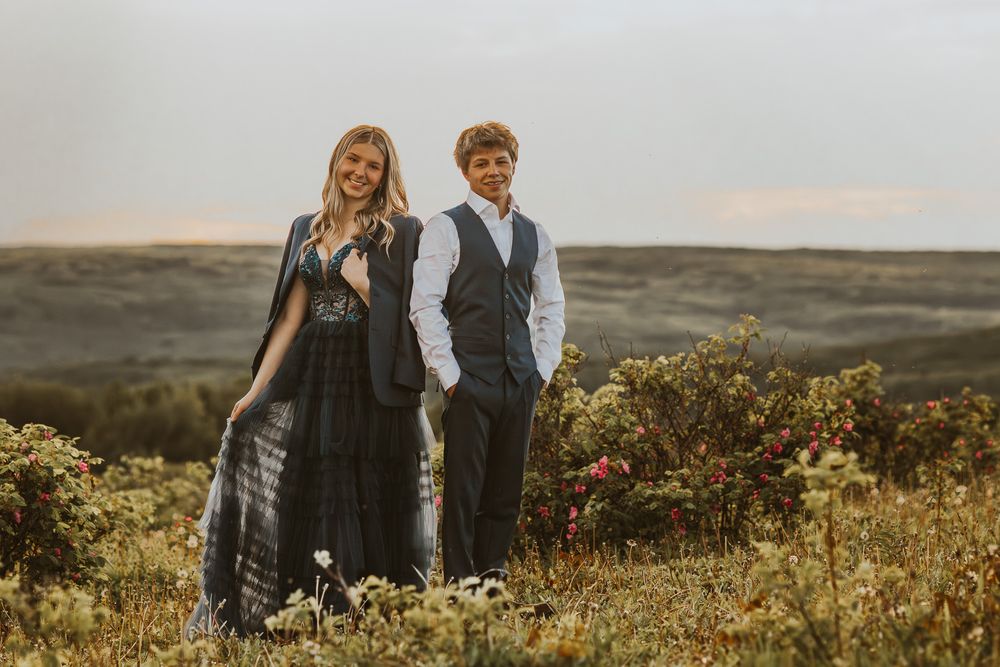 A young woman in a black dress and boy in a vest pose together in a scenic outdoor field at sunset.