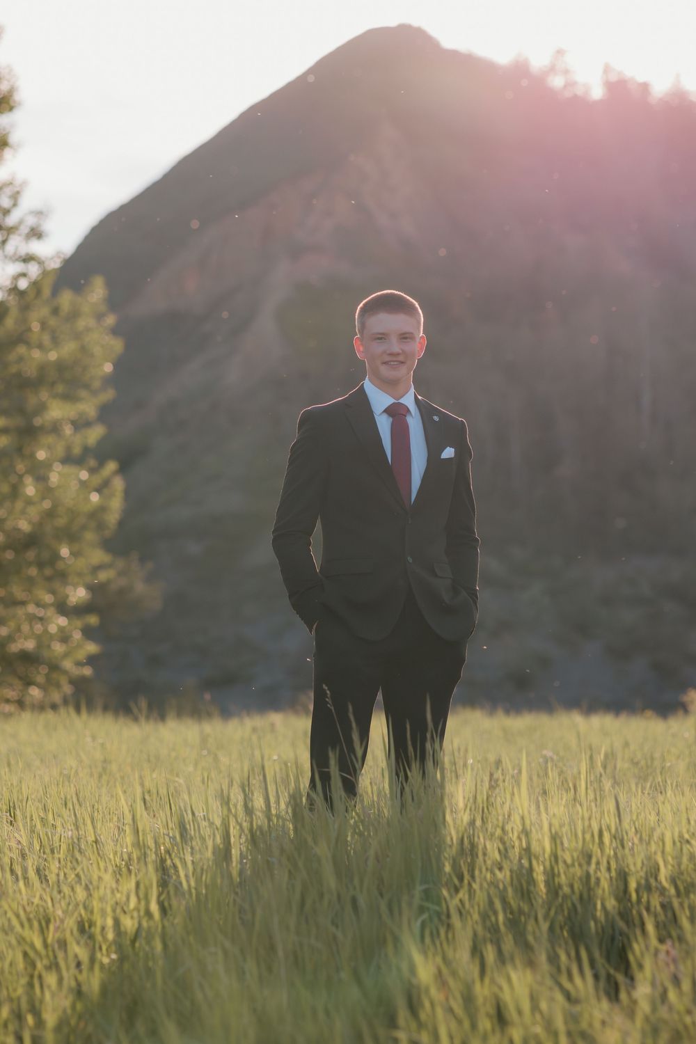 A smiling young man in a black suit standing in a green field with a mountain backdrop.