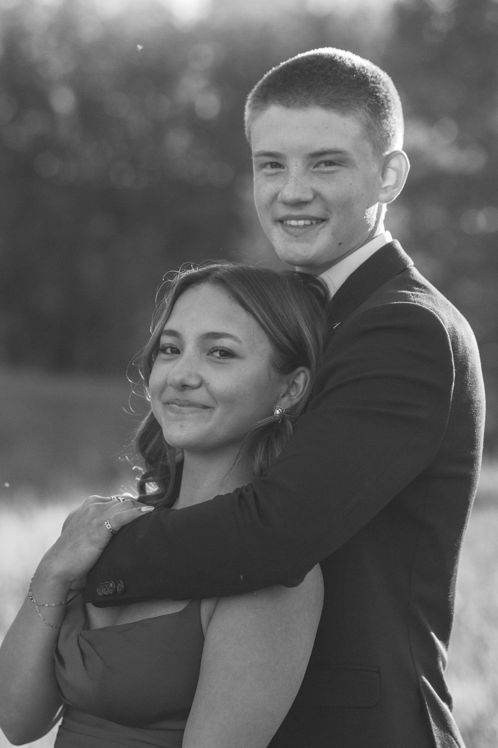 Black and white photo of a young couple in formal wear embracing outdoors, smiling at the camera.