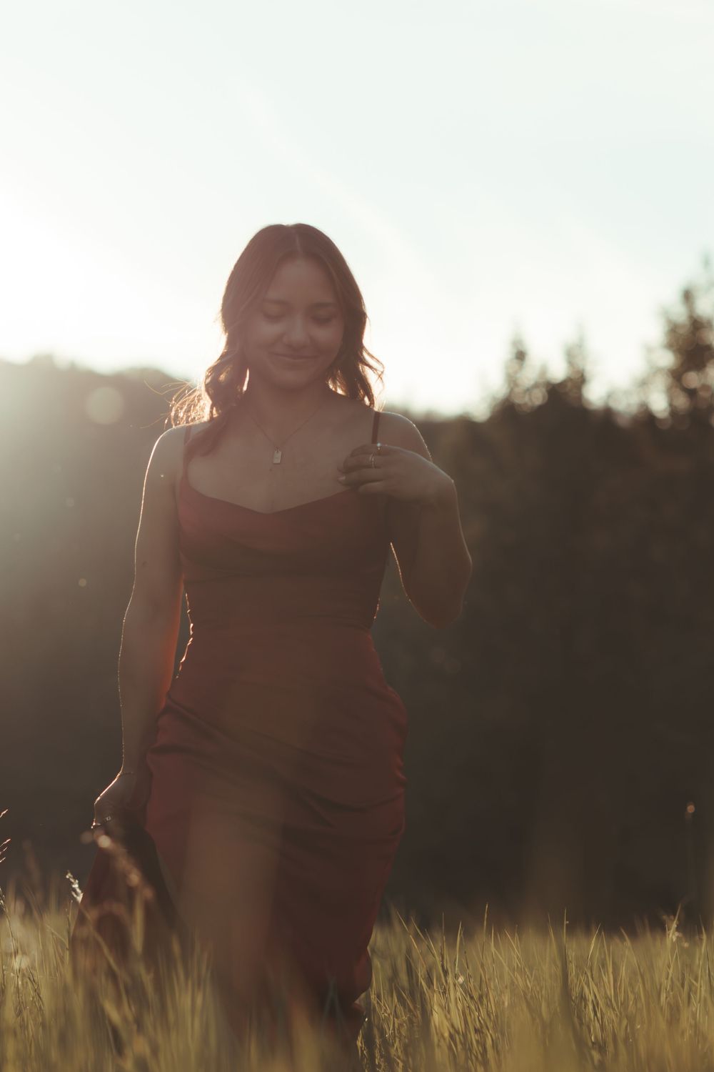 A young woman in a red dress walking through a sunlit grassy field, smiling softly.