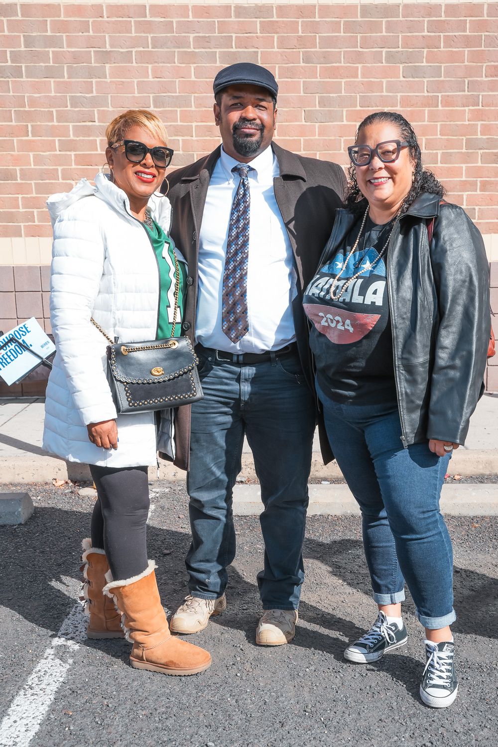Three friends pose together outdoors near a brick wall, dressed in casual fall attire at a political campaign event.