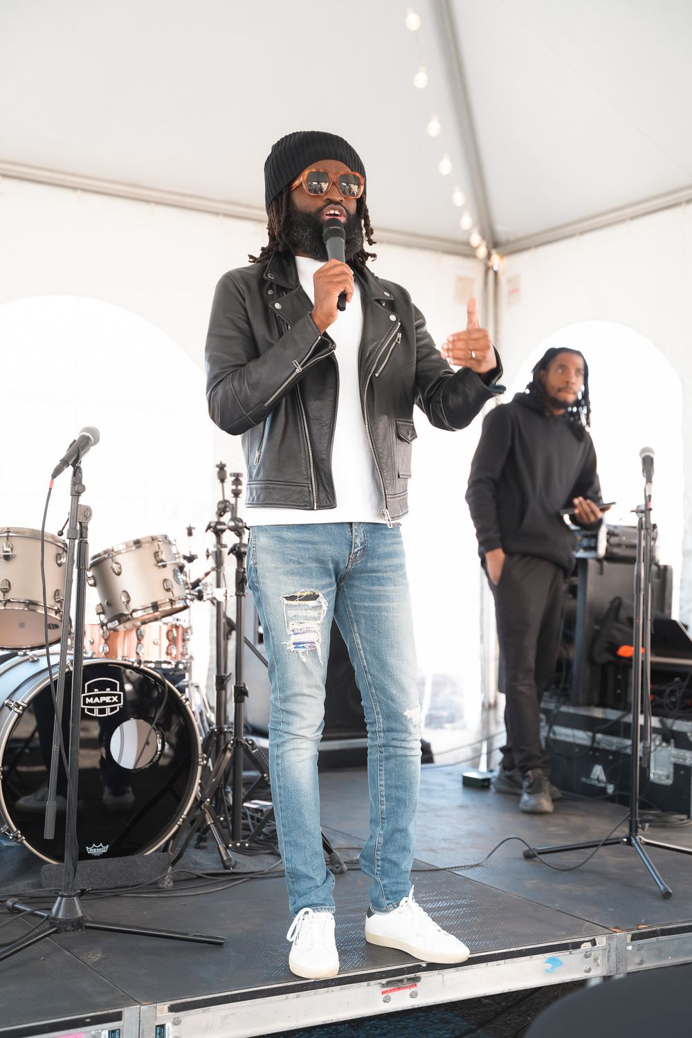 Male performer in black beanie and leather jacket sings on stage with drummer visible in background.
