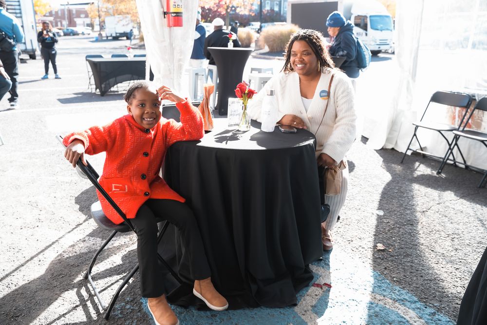 Two women smile together at an outdoor event, seated at a black draped table in autumn.
