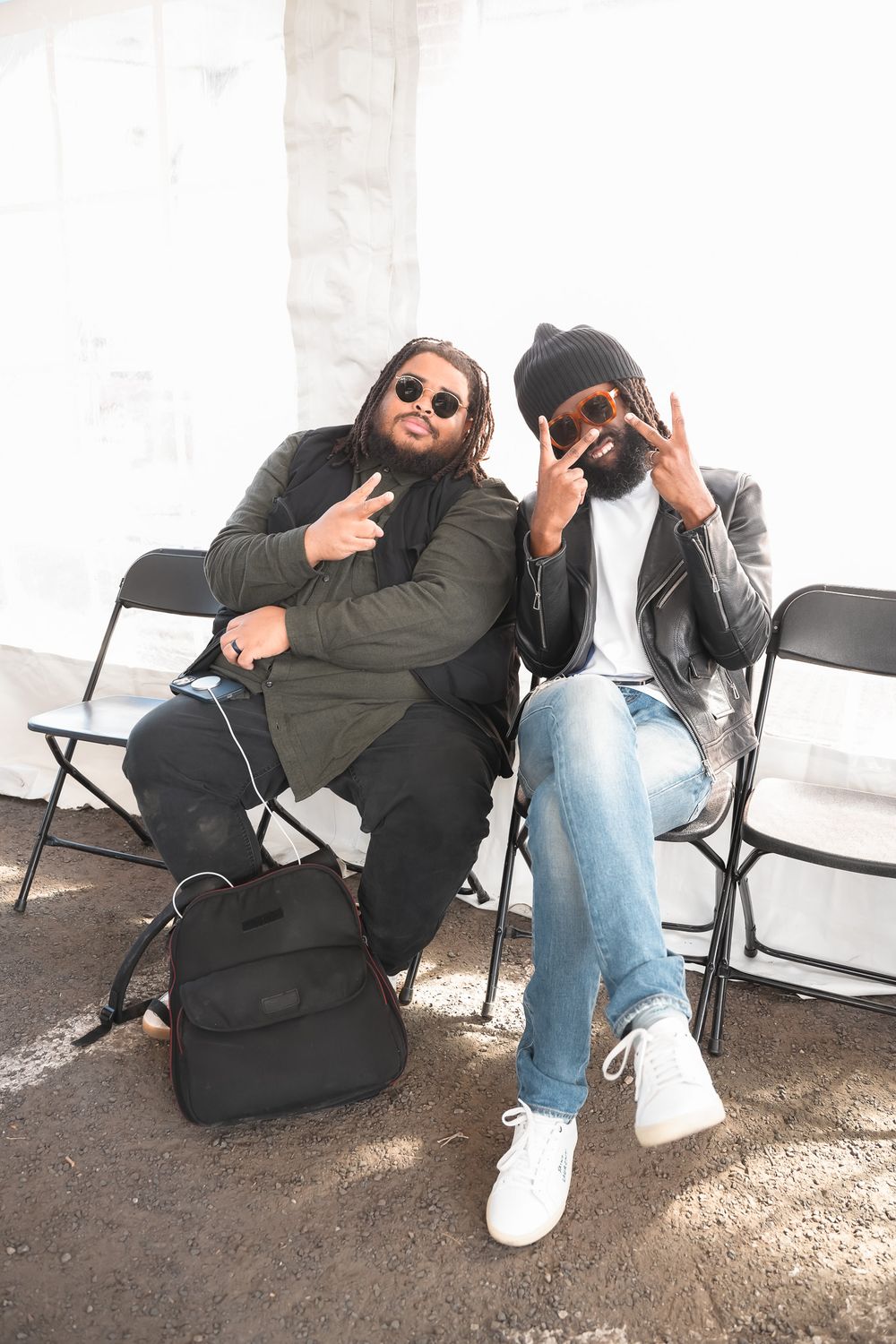 Two Black men sitting in folding chairs making peace signs, wearing sunglasses and casual streetwear outdoors.