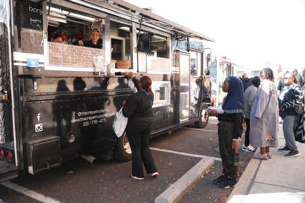 Customers lining up at a black food truck on a sunny day at an outdoor street food event.