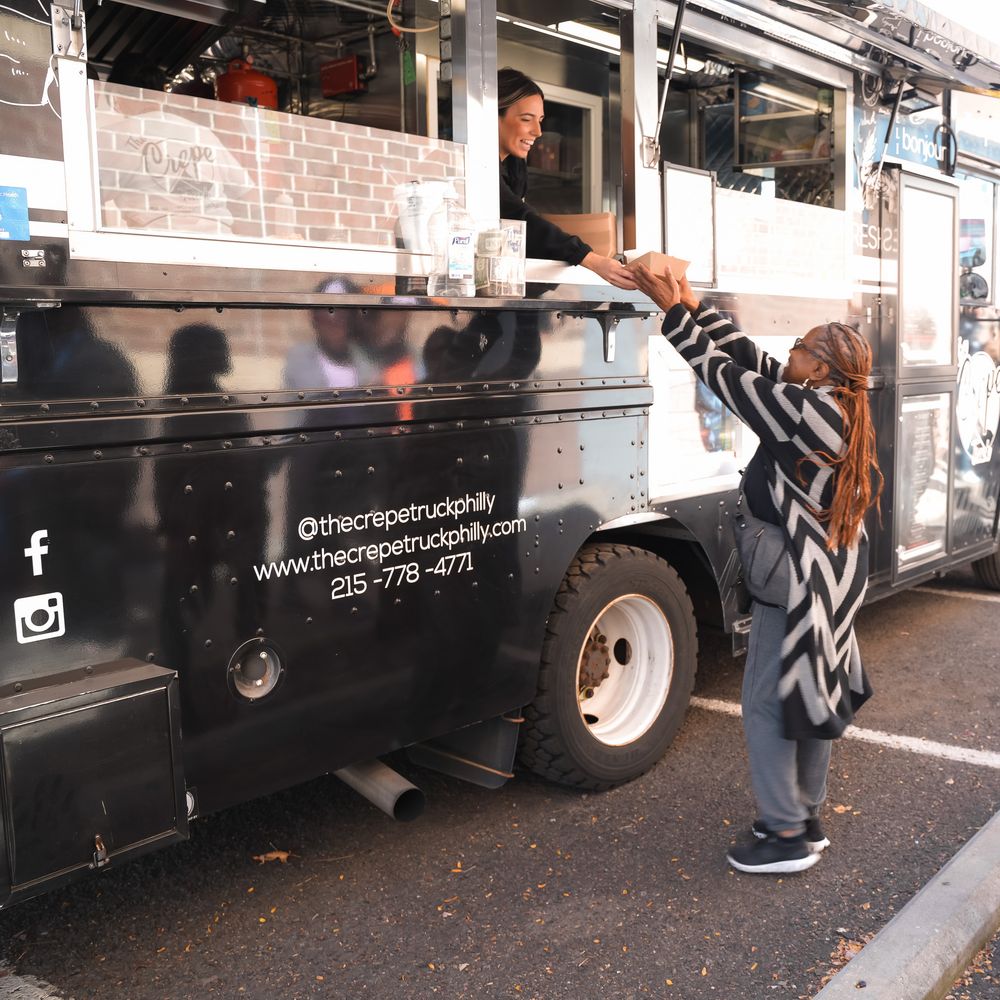 Woman receiving food from a crepe truck vendor at The Crepe Truck Philly food truck in a parking lot.