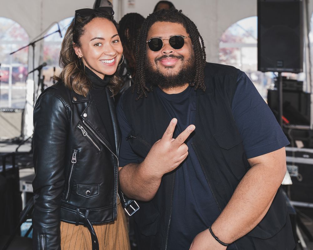 Two friends pose together at an outdoor music event, one wearing a leather jacket and the other flashing a peace sign.