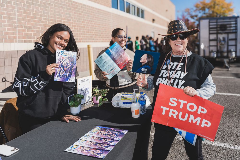 People at outdoor voter registration table holding Stop Trump sign and campaign materials at community event.