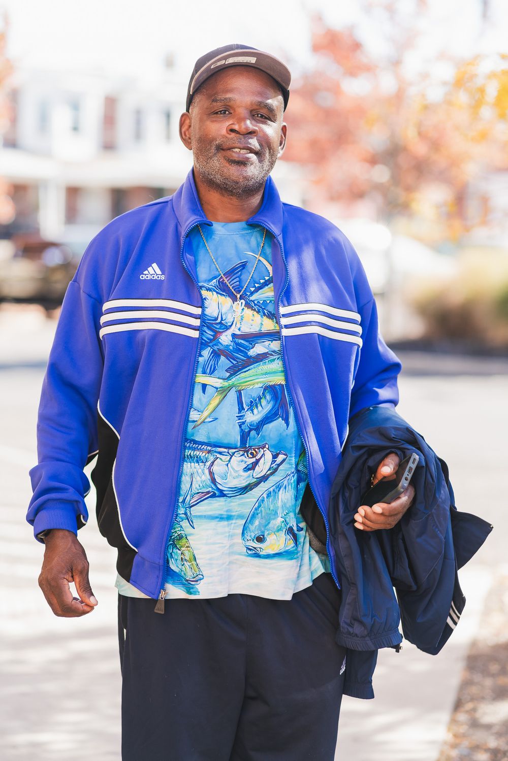 Smiling man in blue Adidas track jacket and fish print shirt walks outdoors on a sunny autumn day.