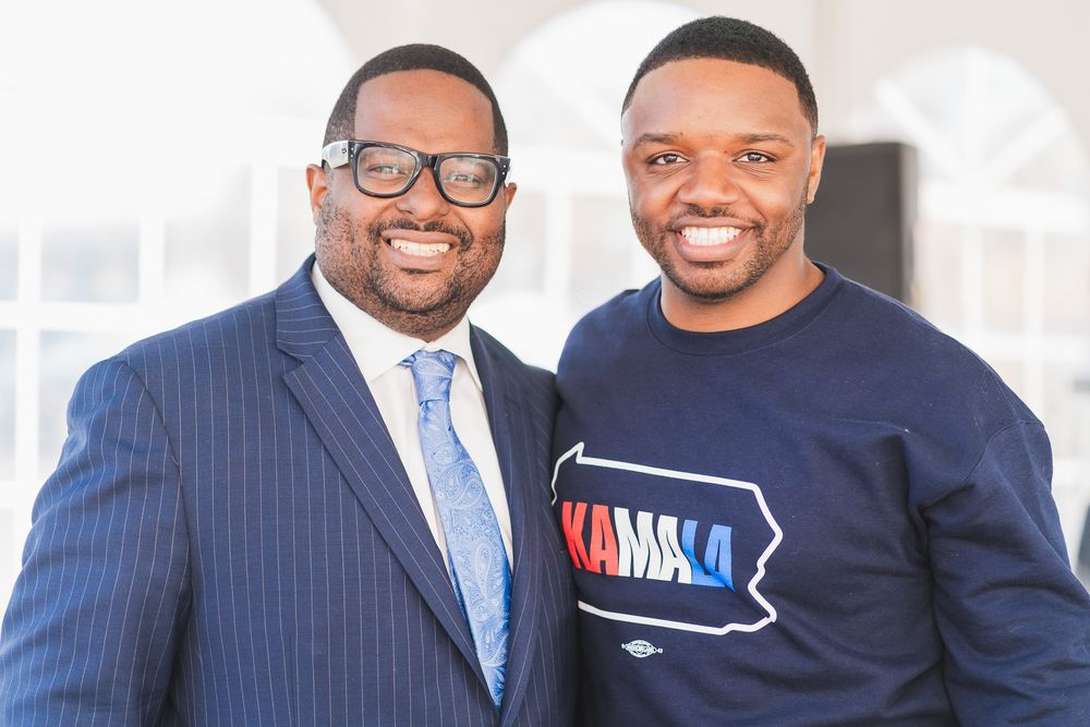 Two Black men smiling together, one in a blue suit and one in a Kamala campaign sweatshirt.