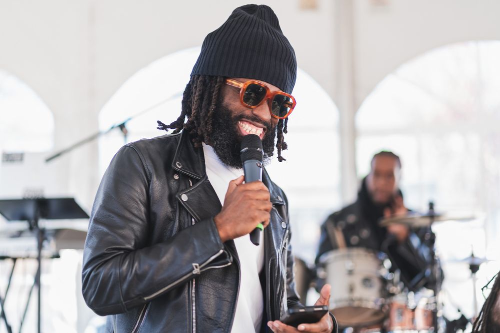 Man with dreadlocks and red sunglasses speaking into microphone at outdoor music event with drummer behind him.
