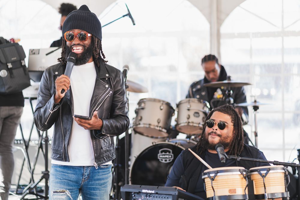 Two musicians perform on stage, one singing into a mic and another playing bongos under a white tent.