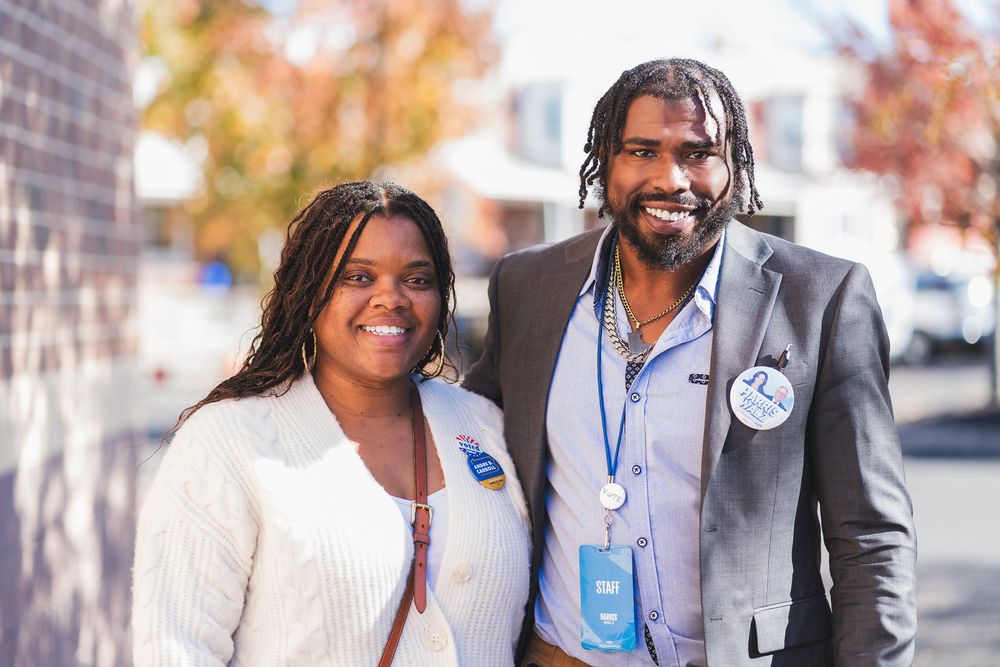 Two smiling people wearing name badges pose outdoors on a sunny autumn day.