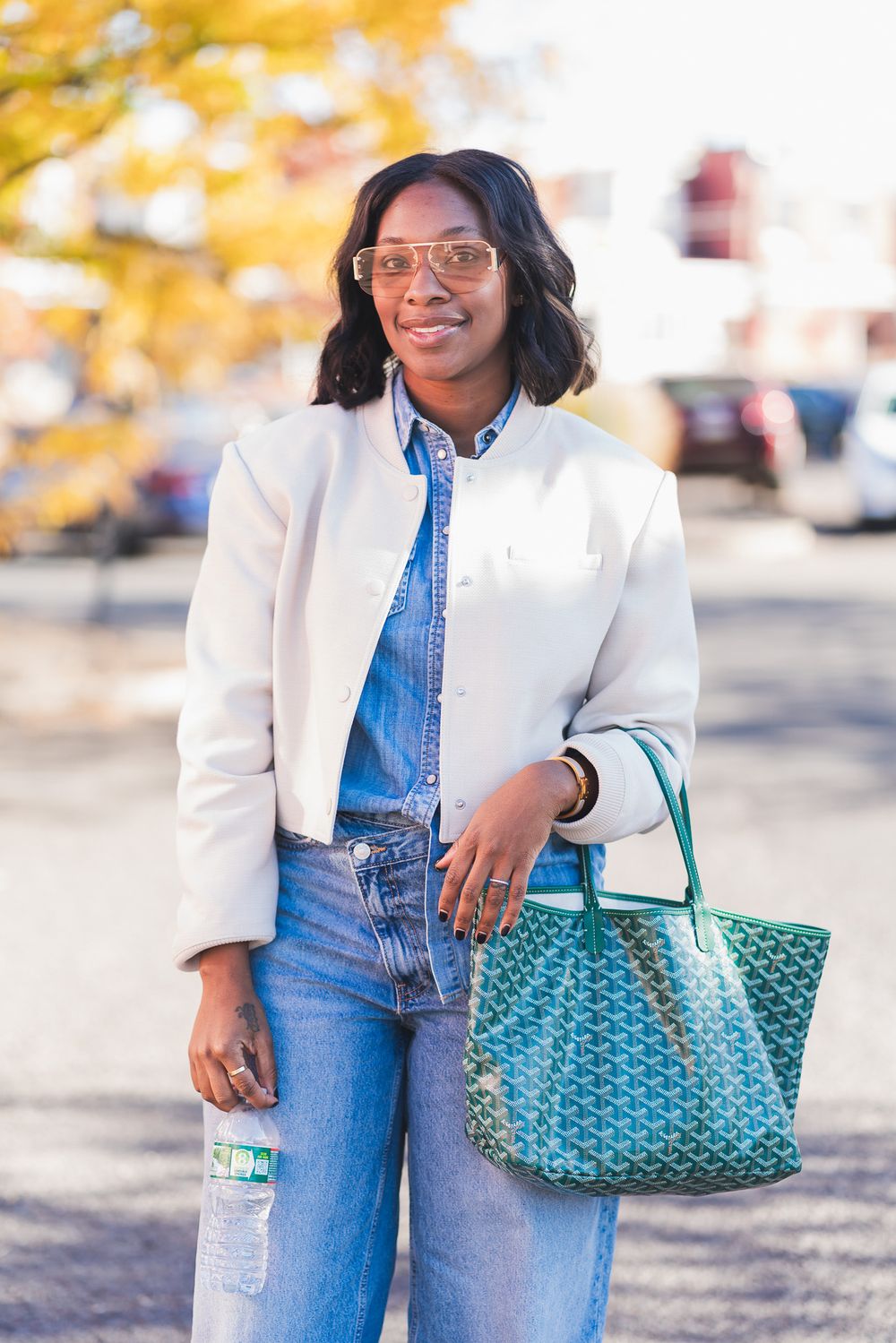 Stylish Black woman in denim outfit, white blazer, and gold sunglasses holding a green Goyard tote bag outdoors.