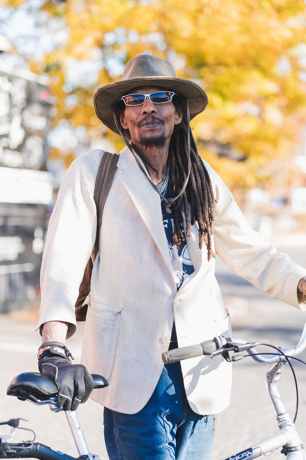 Man with dreadlocks, wide-brim hat, and sunglasses standing with a bicycle on a sunny autumn street.