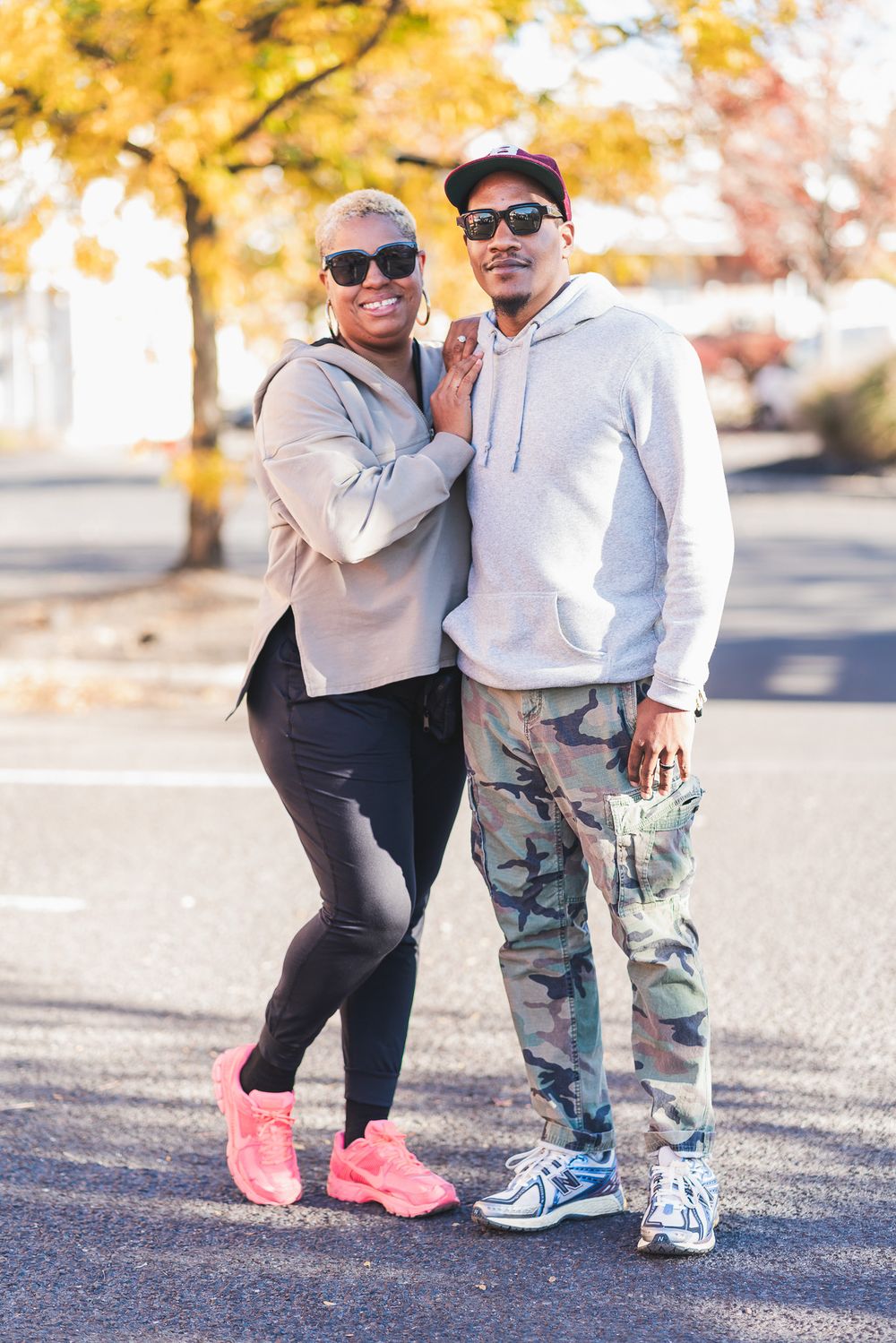 Black couple posing outdoors in casual streetwear, woman in pink sneakers and man in camo pants and gray hoodie.
