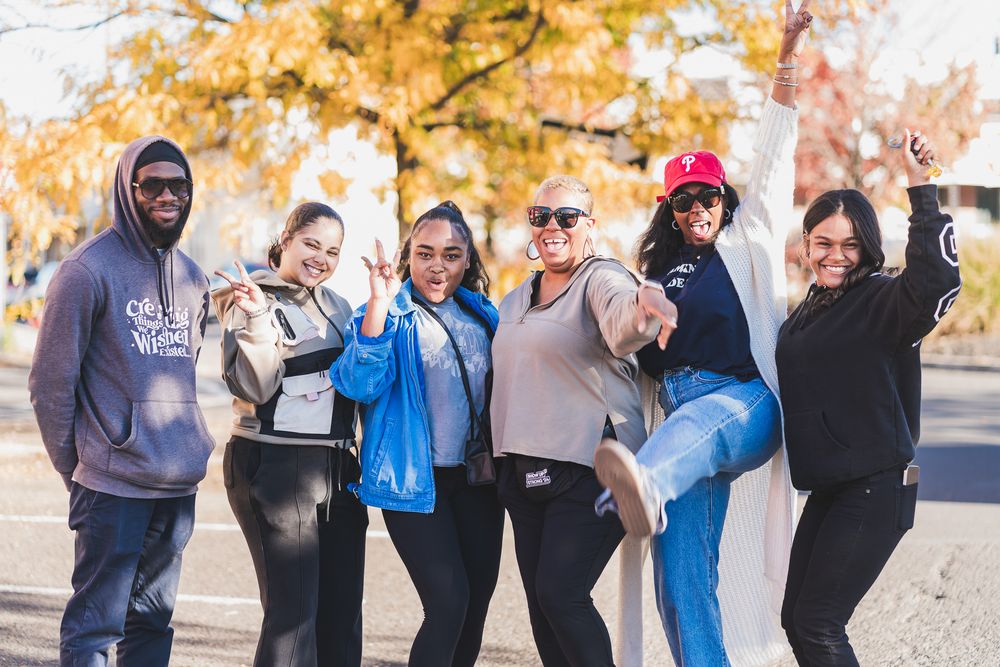 A diverse group of six friends posing and smiling outdoors in fall, wearing casual clothes near colorful autumn trees.