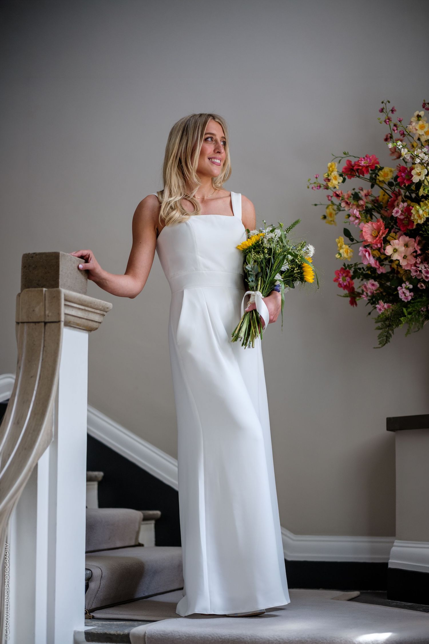 Bride in elegant white sleeveless gown holding colorful bouquet on staircase beside floral arrangement.