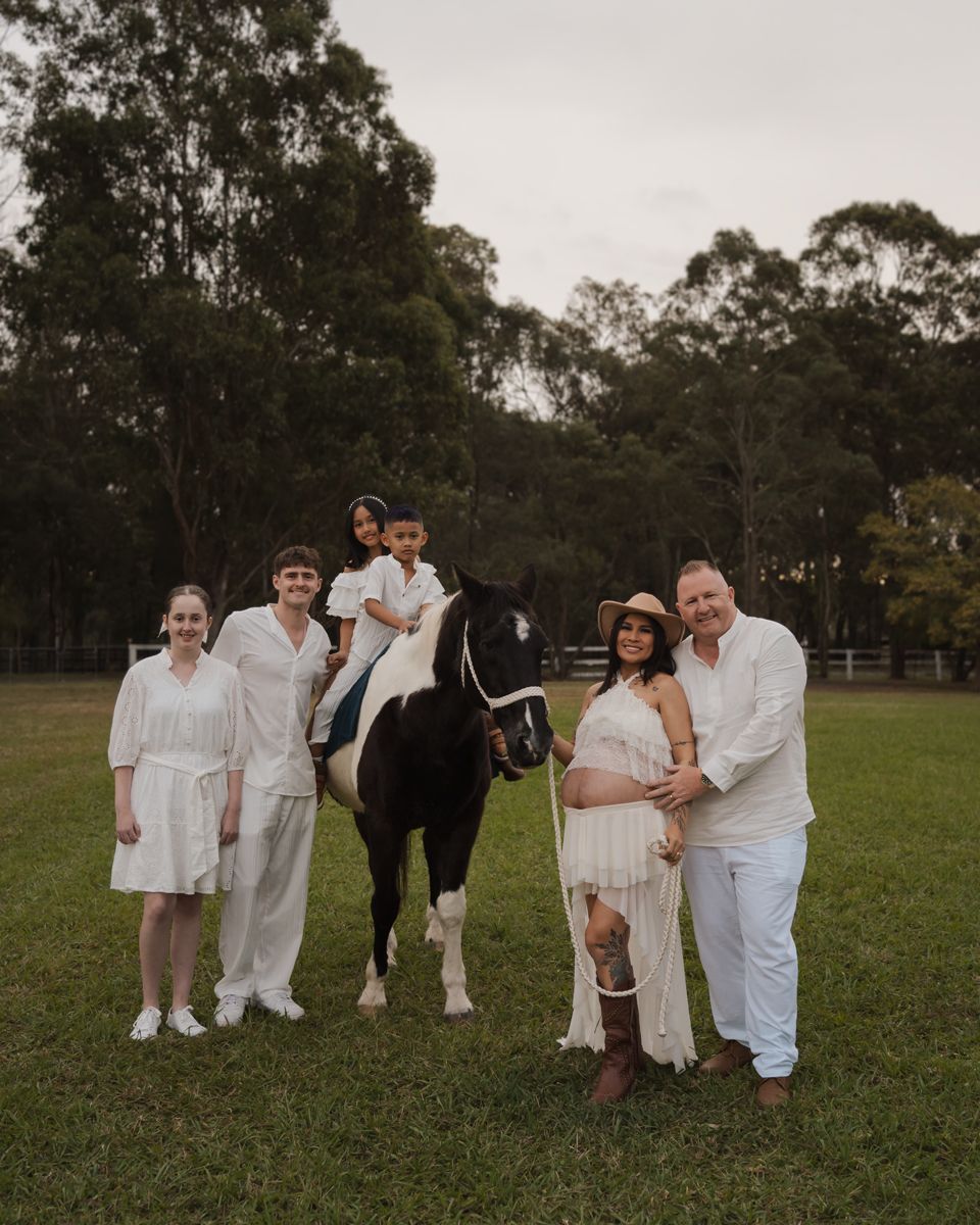 Family in white outfits posing with a black and white horse on a green field, with a child sitting on the horse in sydney photoshoot