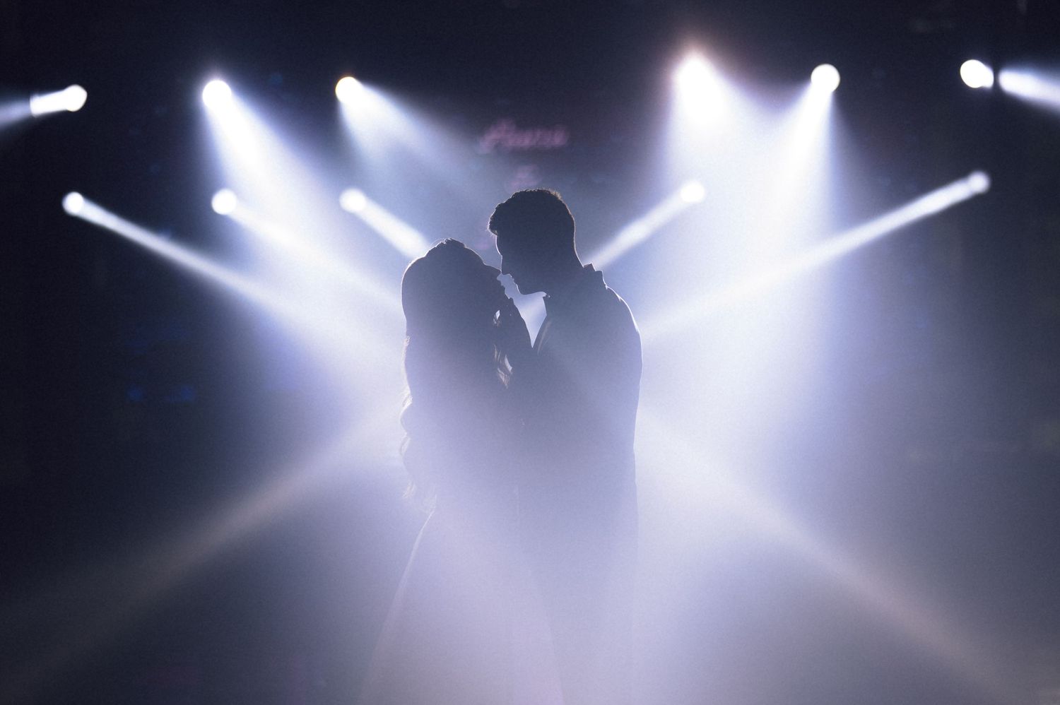 Silhouette of a couple sharing a romantic moment on stage, surrounded by dramatic white spotlights.