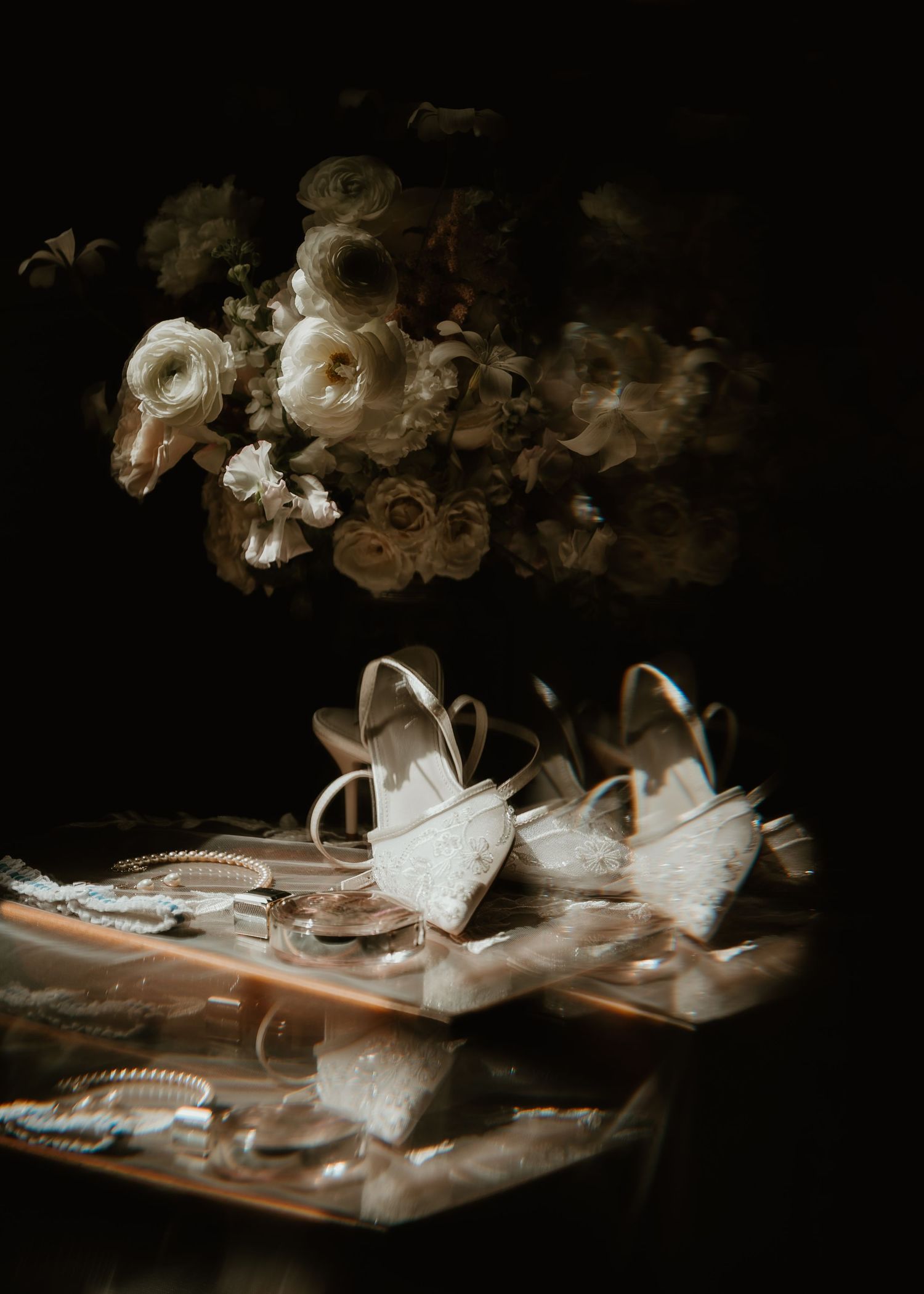 Elegant bridal flat lay with lace heels, gold rings, and white floral bouquet on dark moody background.